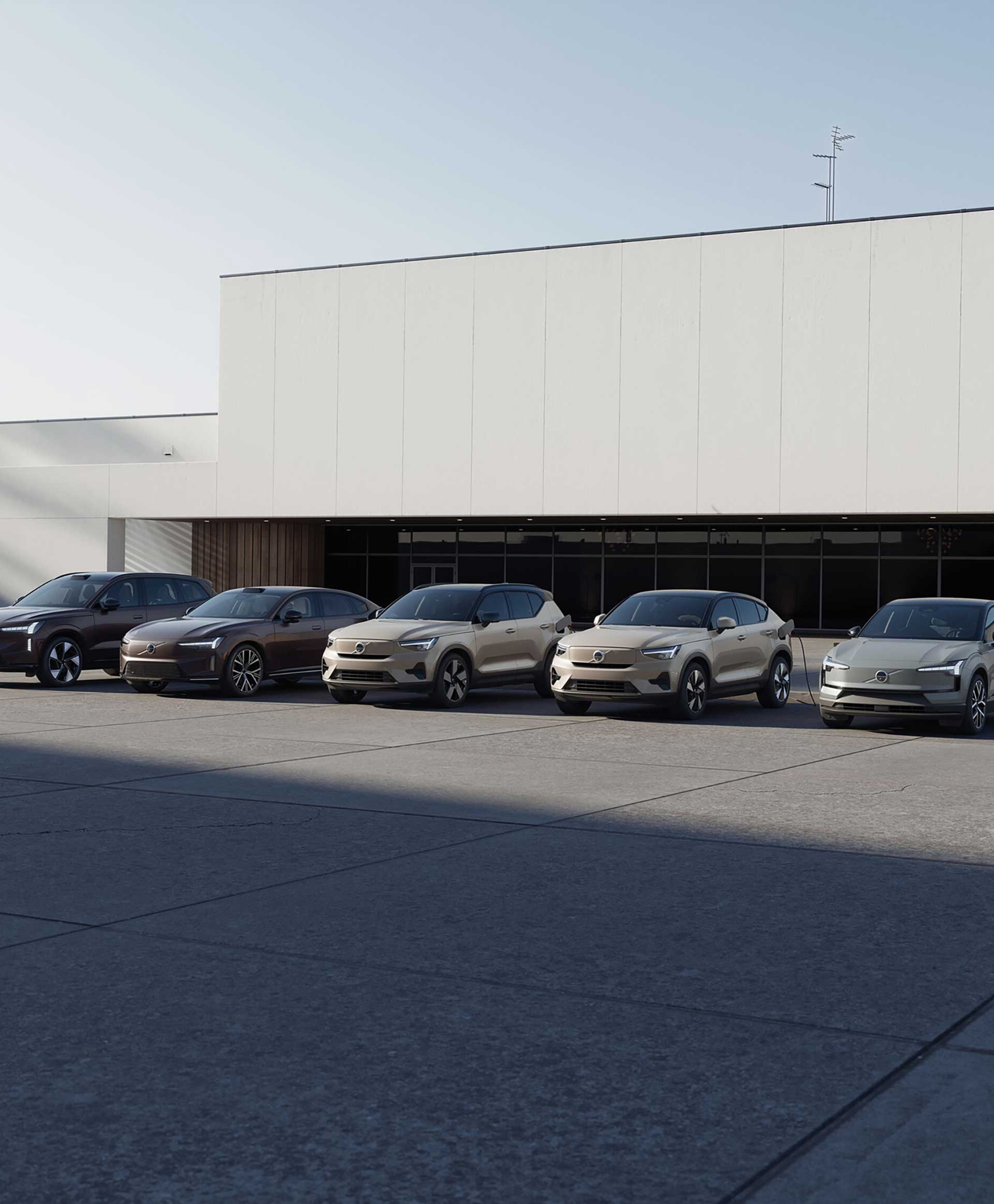 En række på fem stilfulde, moderne Volvo elbiler i dæmpede farver er parkeret foran en minimalistisk hvid bygning. Scenen er rolig og ordnet under en klar himmel.