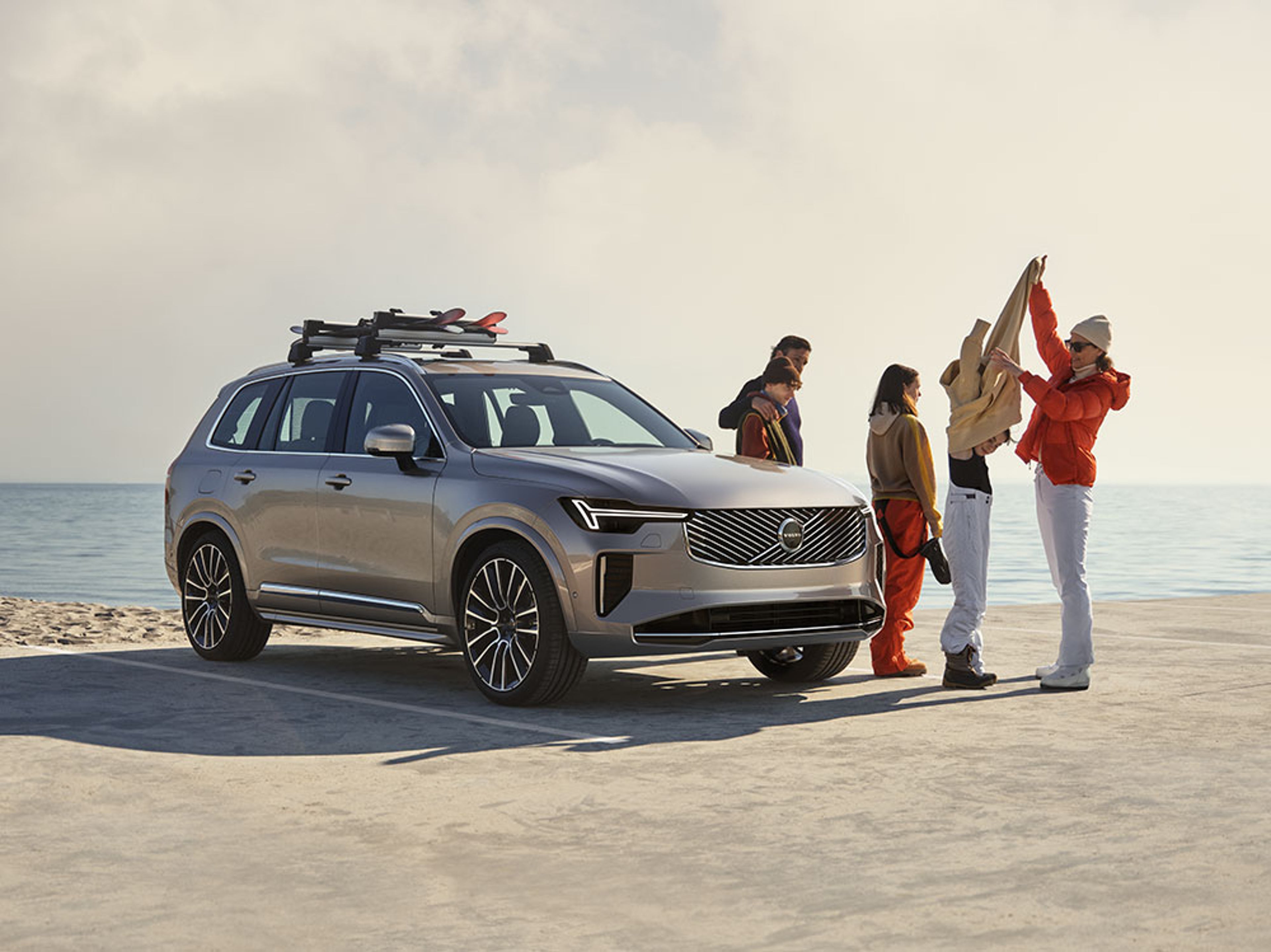 XC90 Argent Crépuscule vu de l’avant garé sur un parking près d’une plage avec une famille de 4 personnes