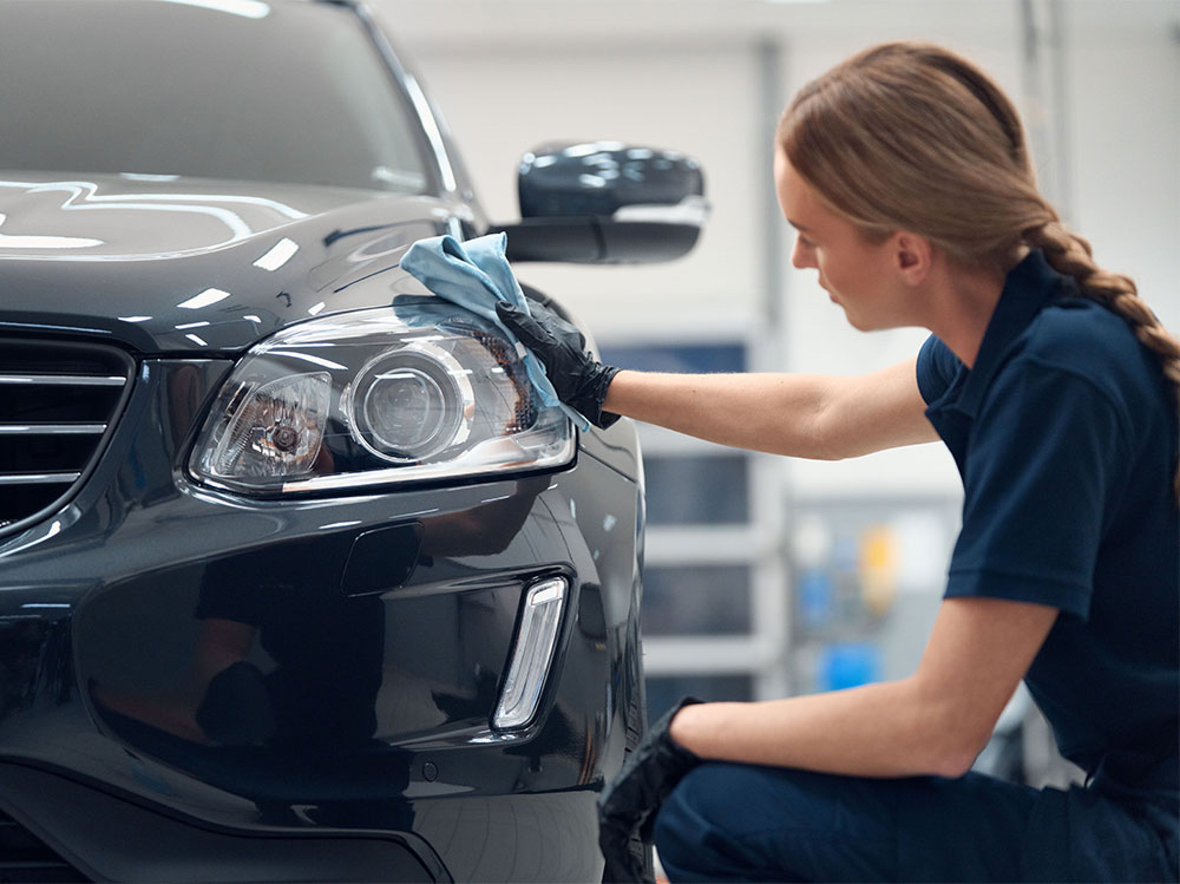 Femme nettoyant le phare d’un Volvo noir avec un chiffon bleu dans un garage.