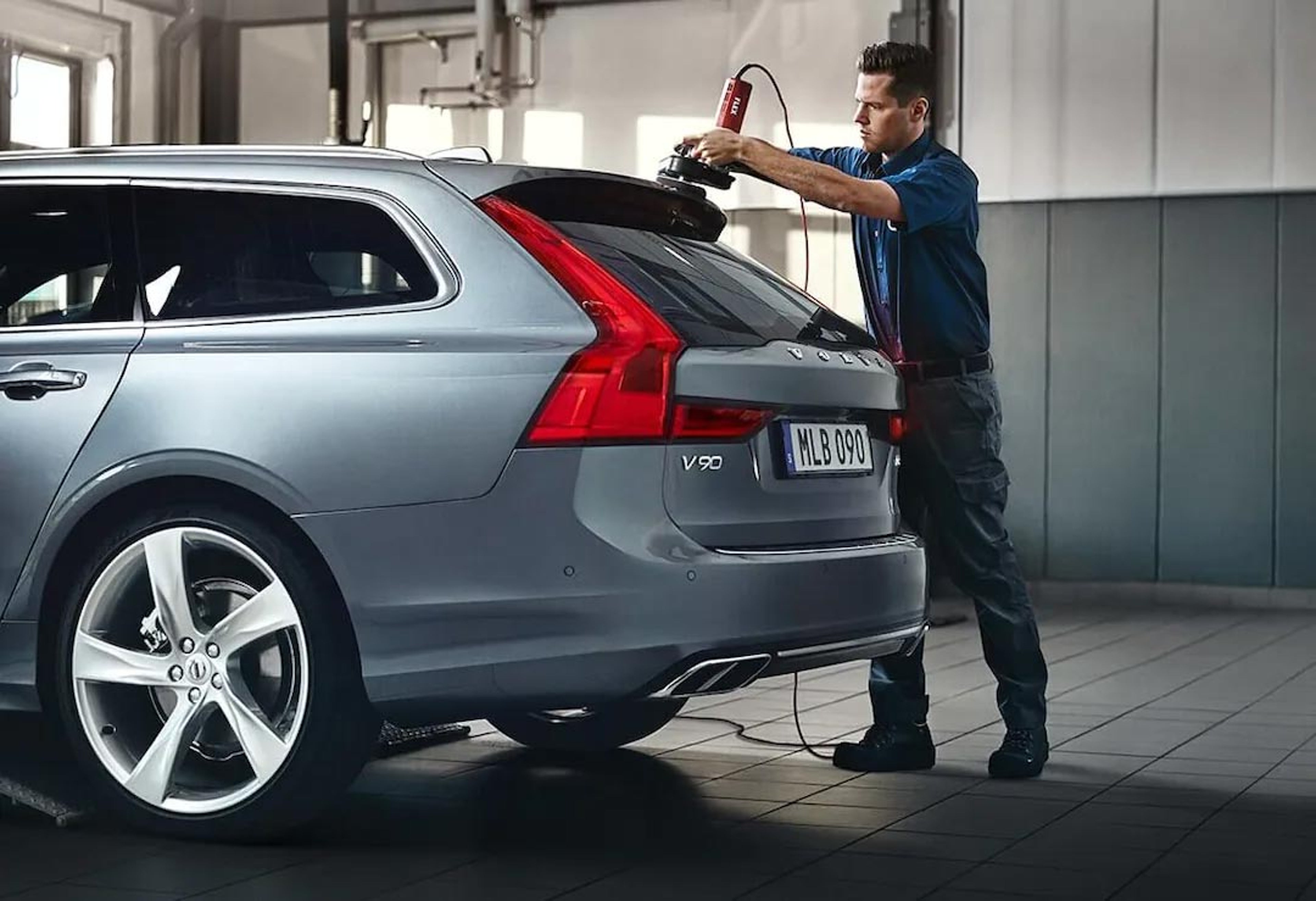 Ein Volvo Personal Service Techniker steht am Heck eines Volvo V90 in einer Werkstatt und bearbeitet den Lack des Fahrzeugs mit einer Poliermaschine.