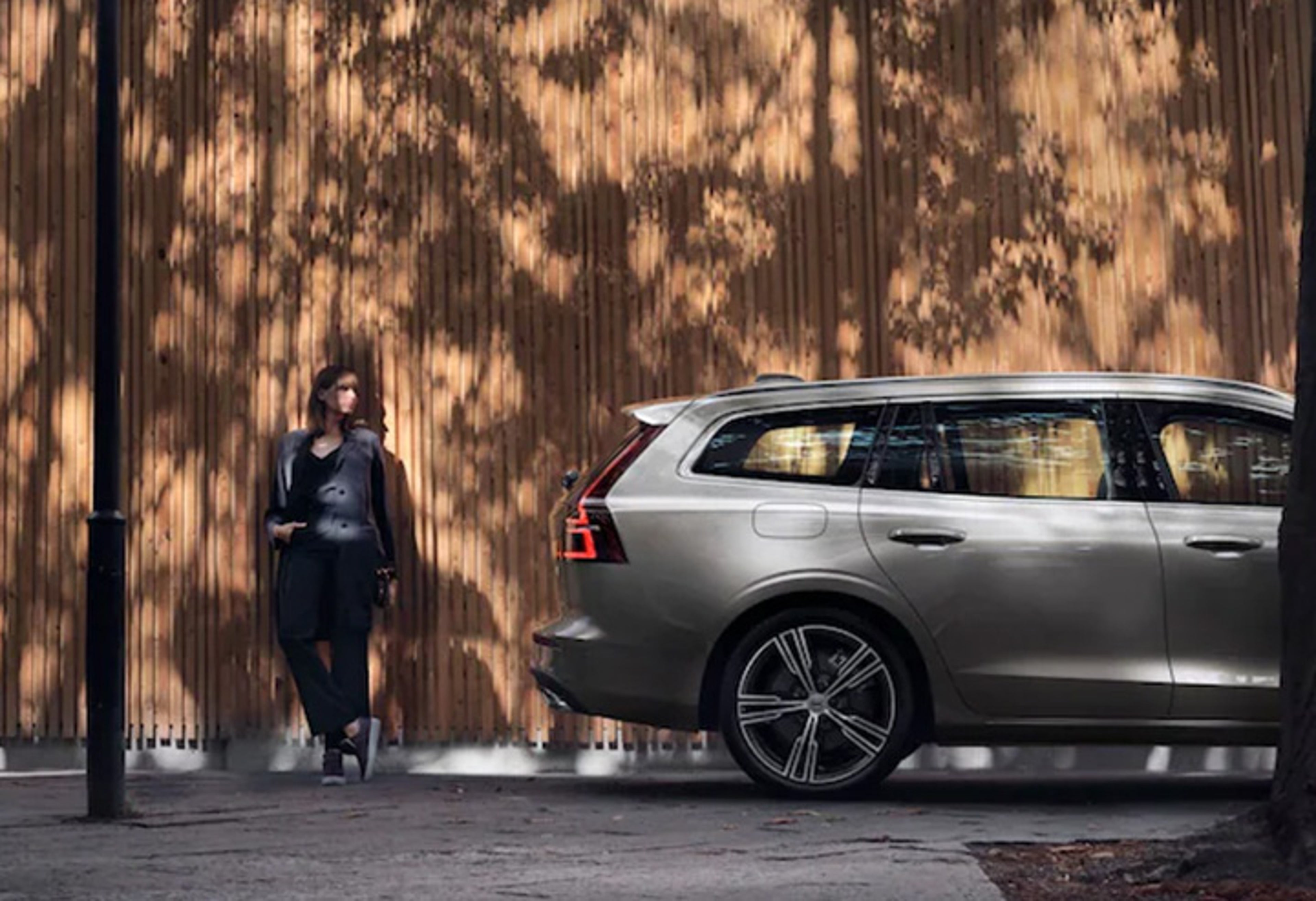 Eine Frau steht hinter einem silbernen Volvo V90 Kombi und lehnt an einer Holzwand, auf der Schatten von Bäumen fallen.