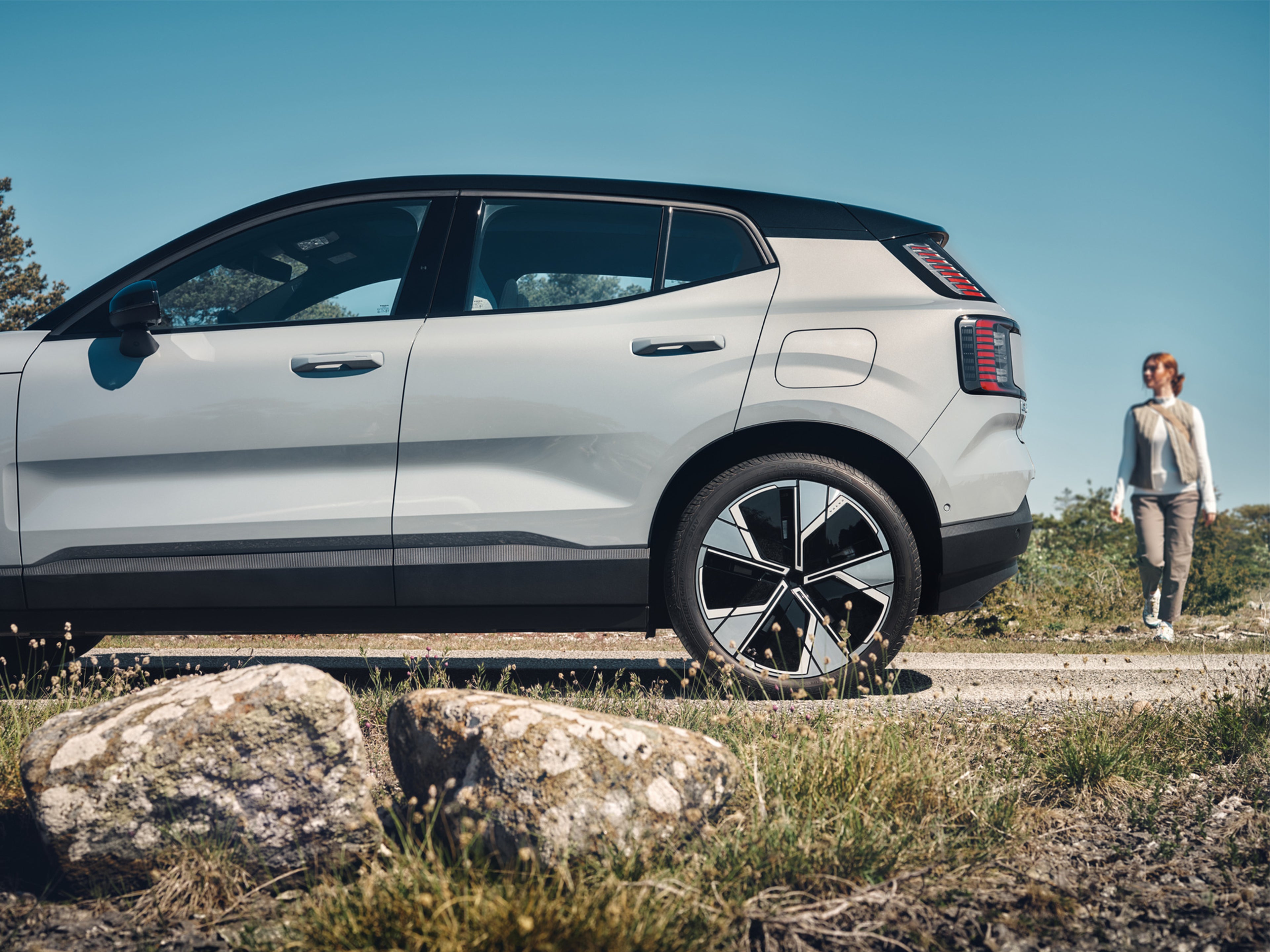 Nainen kävelee kohti metsätielle pysäköityä Vapor Grey -väristä Volvo EX30 täyssähköautoa.