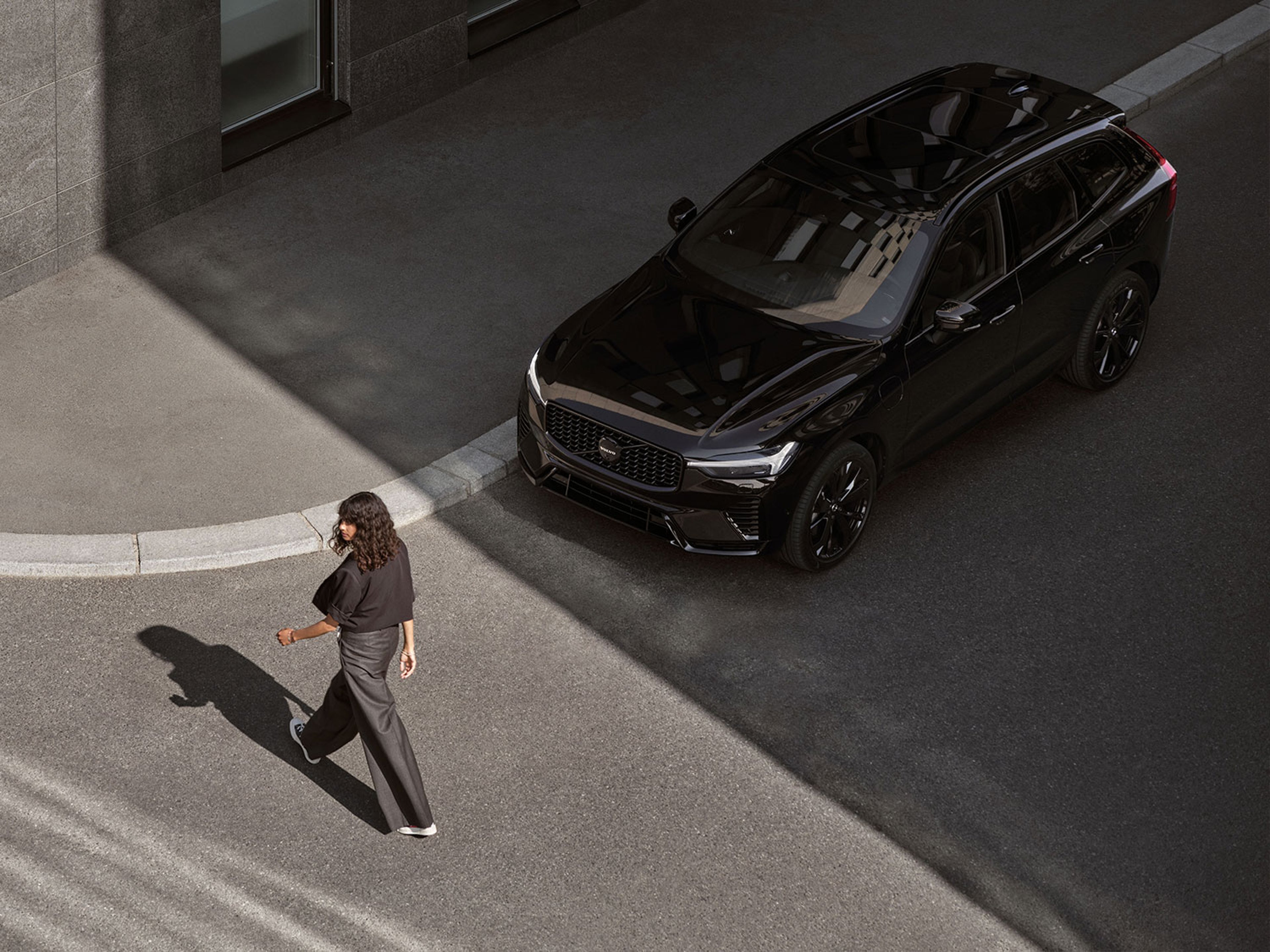 Une Volvo XC60 noire garée dans la rue, avec une femme qui passe devant. Vue de dessus.