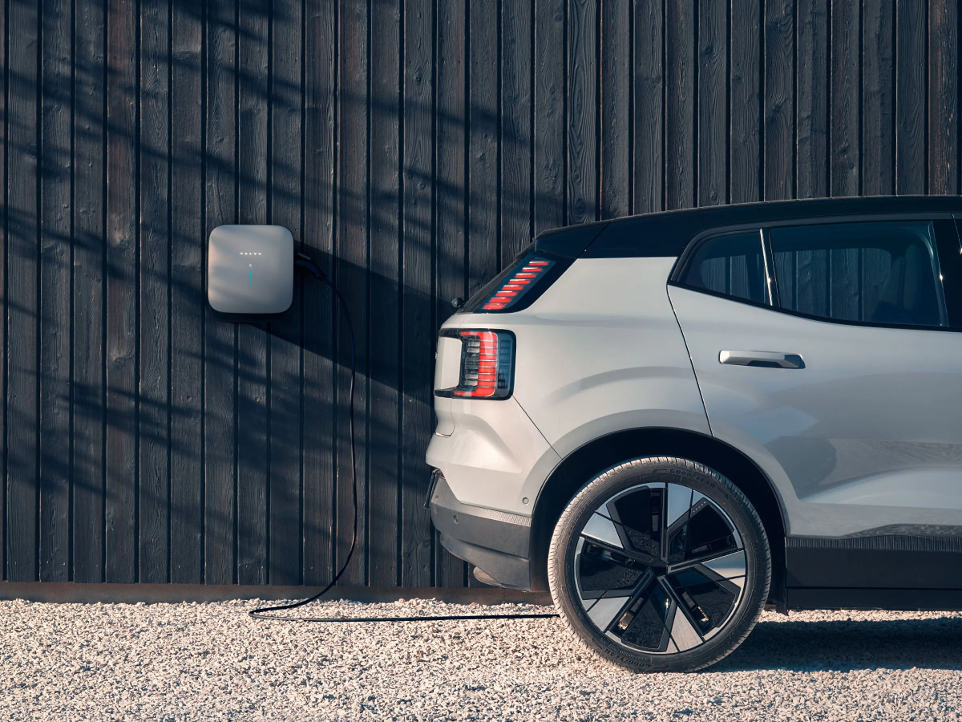 Une Volvo EX30 gris argenté avec un toit noir se recharge à une borne de recharge fixée à un mur en bois sombre.