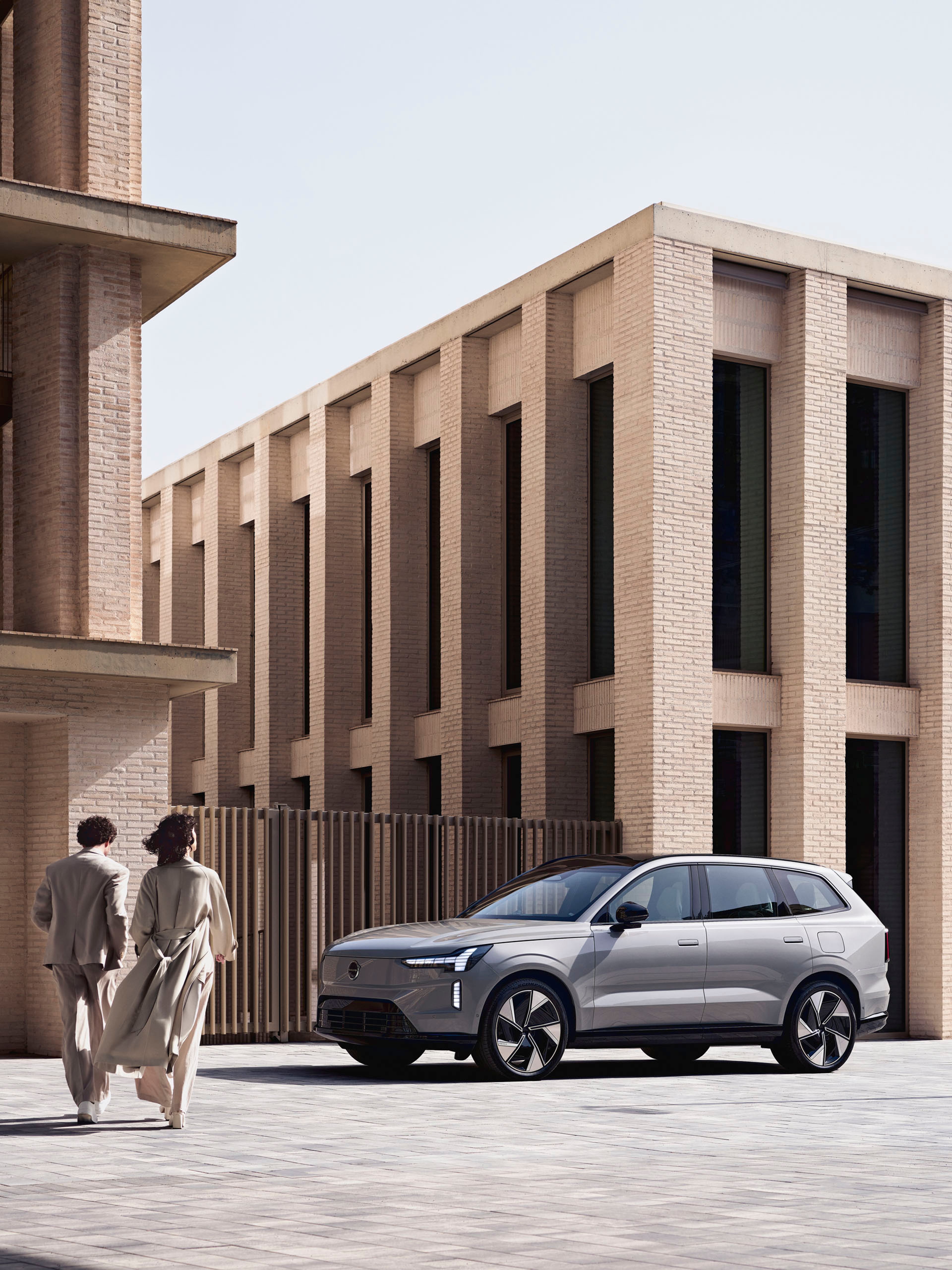 Dos personas caminando hacia un Volvo EX90 eléctrico aparcado entre edificios.