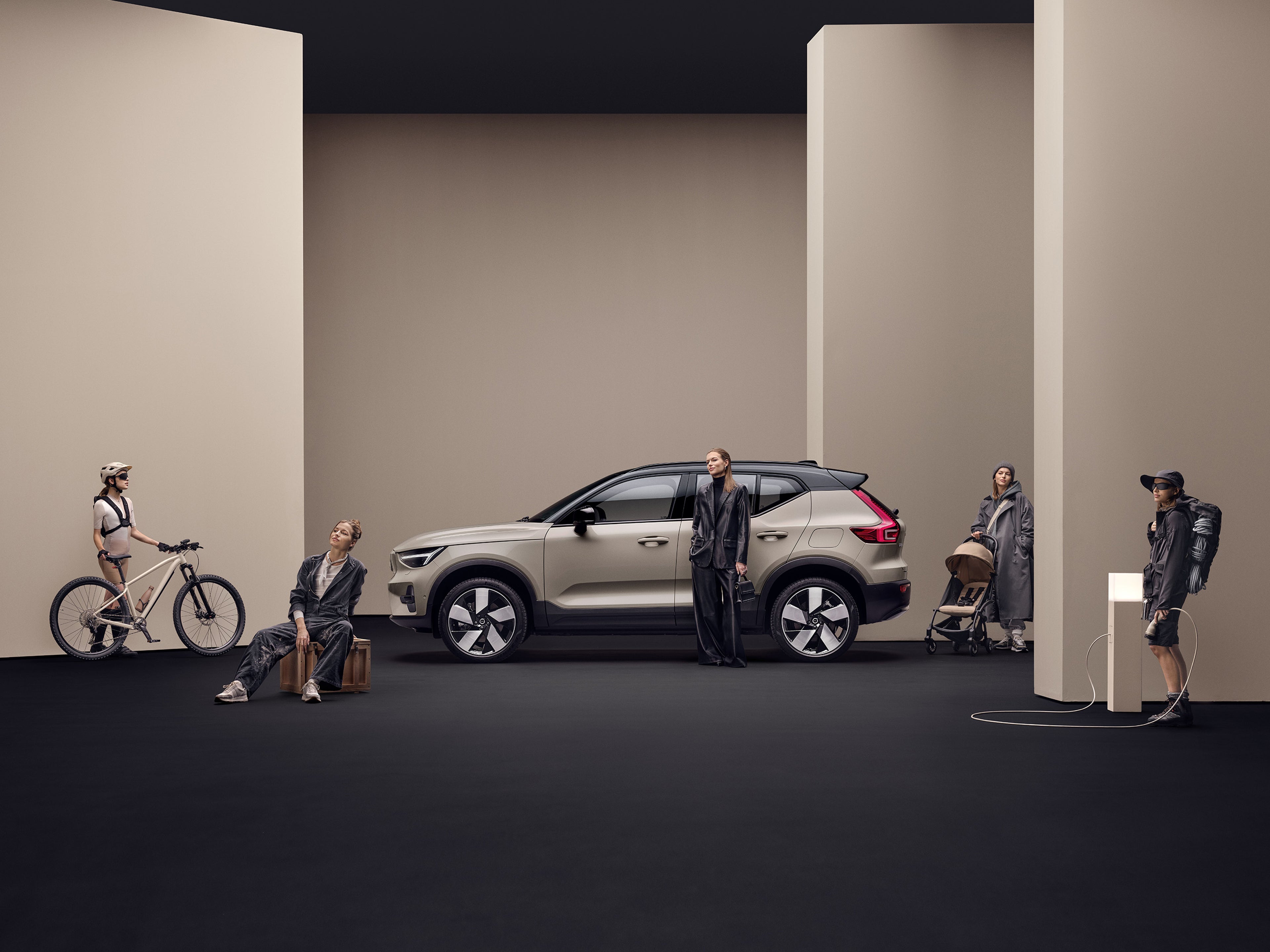 A side profile image of the Volvo EX40 in colour Sand Dune. The car is surrounded by various people who are in all different stages of life, one with a child buggy, one hiking, one is a stylish piece and then one with a bike. This highlights that the EX40 is for every you.