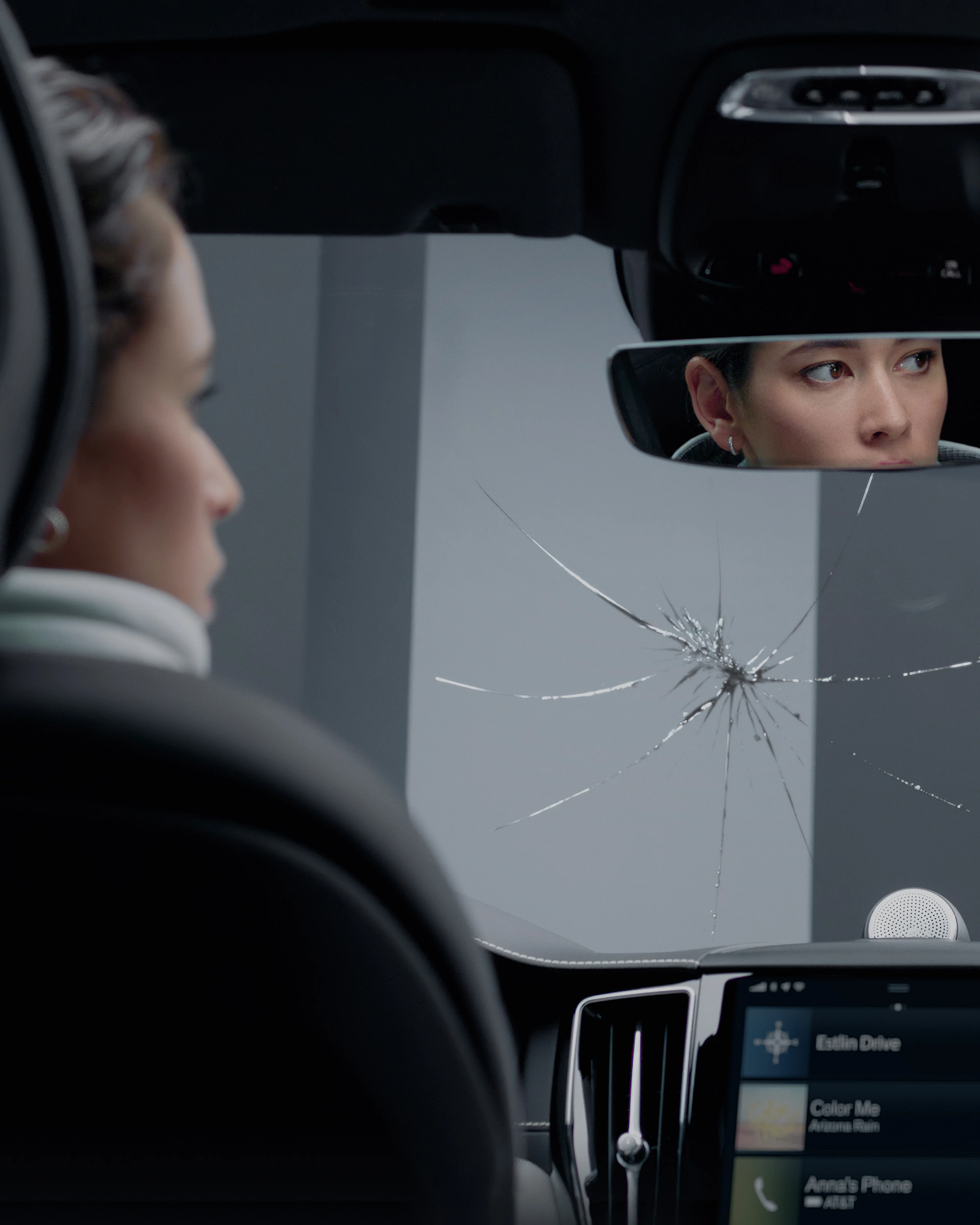 Une femme est vue de dos dans une voiture, avec un pare-brise fissuré et son reflet dans le rétroviseur.