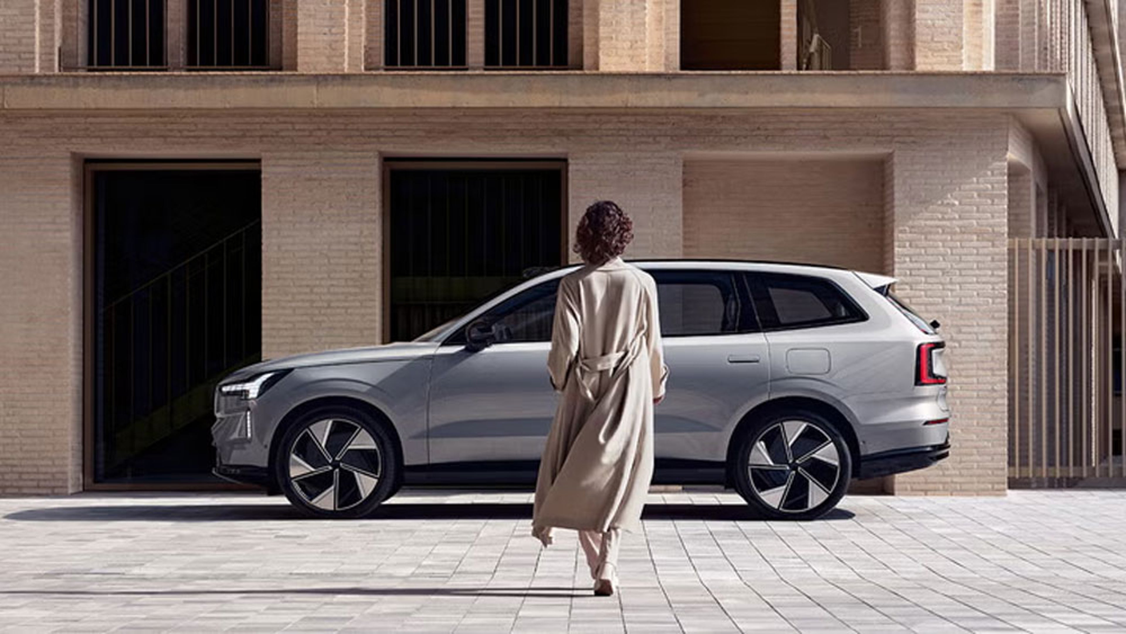 Femme en manteau beige marchant vers un EX90 argenté garé devant un bâtiment moderne en briques