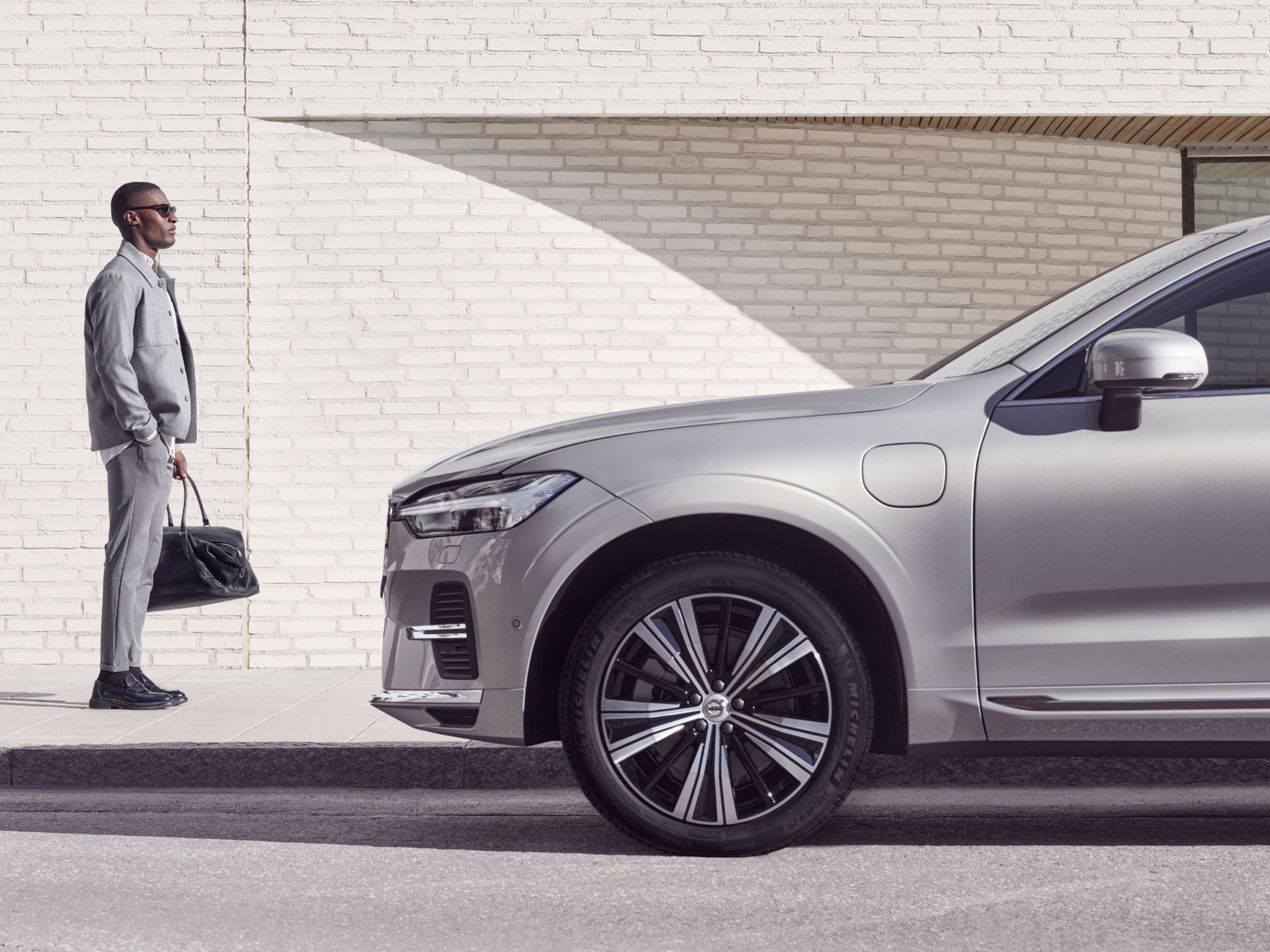 Ein grau-gekleideter Mann mit Tasche steht vor einem parkenden, silbernen Volvo XC90.