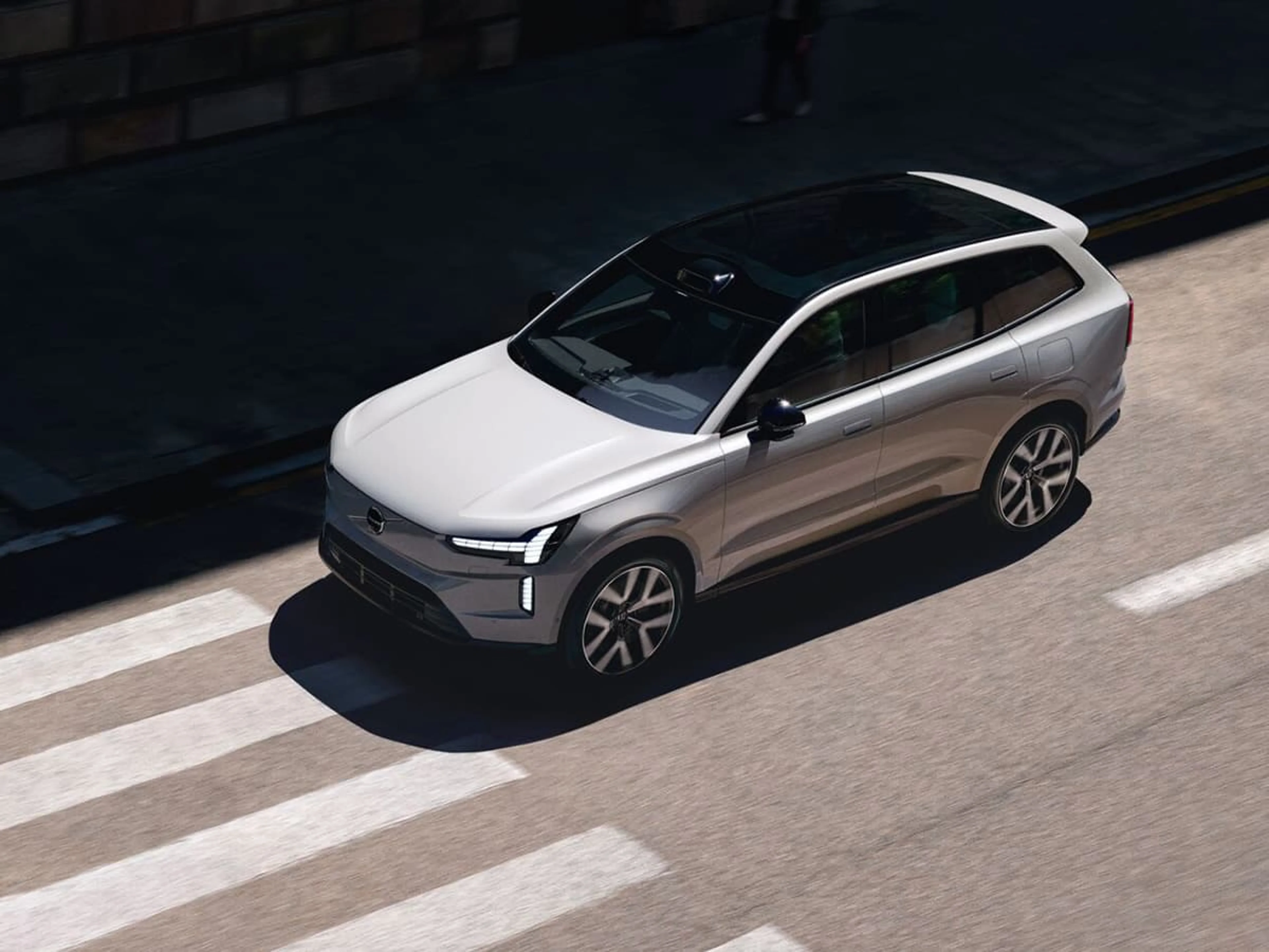 Een zilvergrijze Volvo EX90 elektrische SUV rijdt onder op de weg, vogelperspectief.