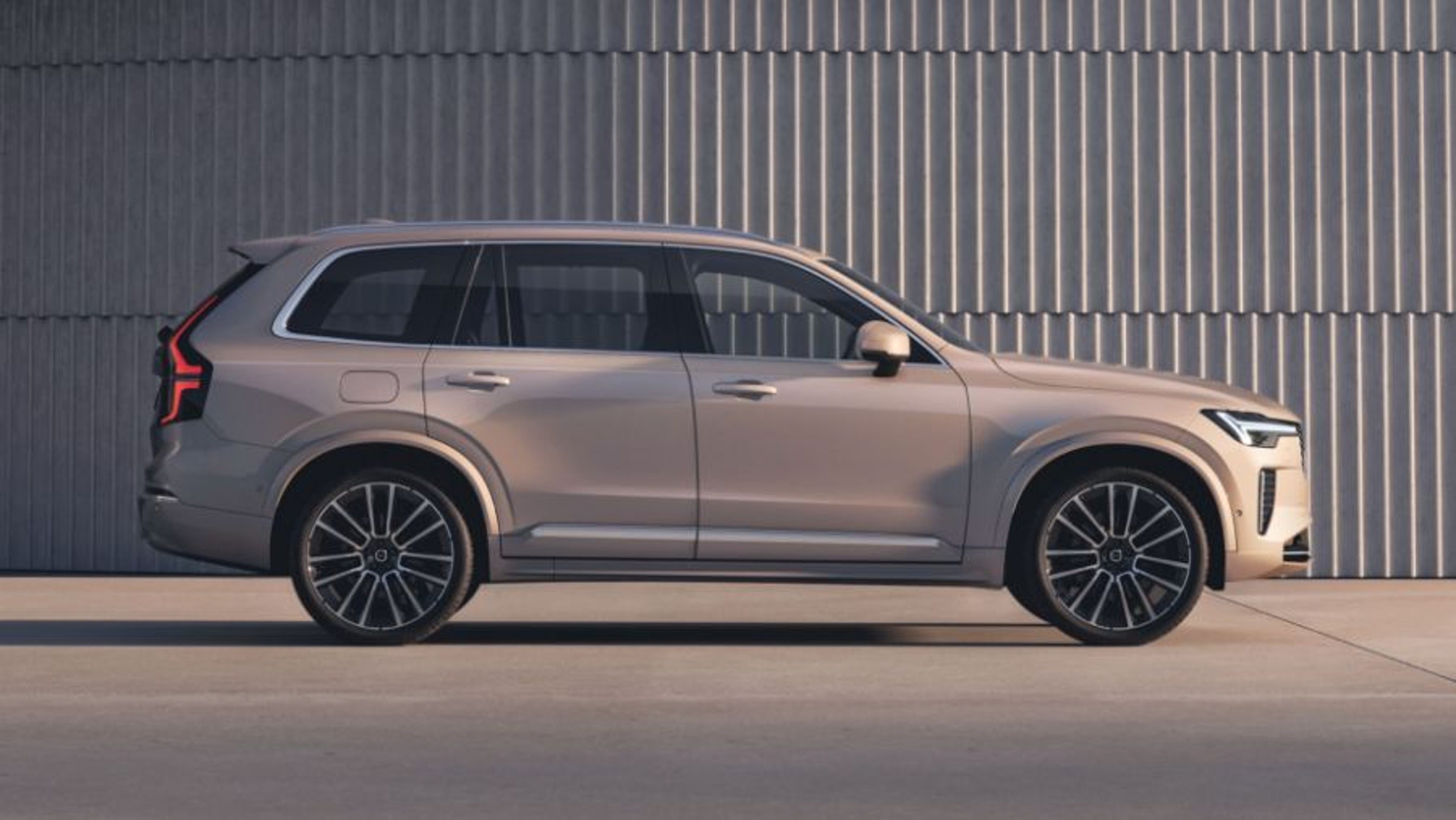 Srebrni Volvo XC90 prikazan iz bočnega profila, parkiran pred kovinsko rebrasto steno v studijskem okolju.