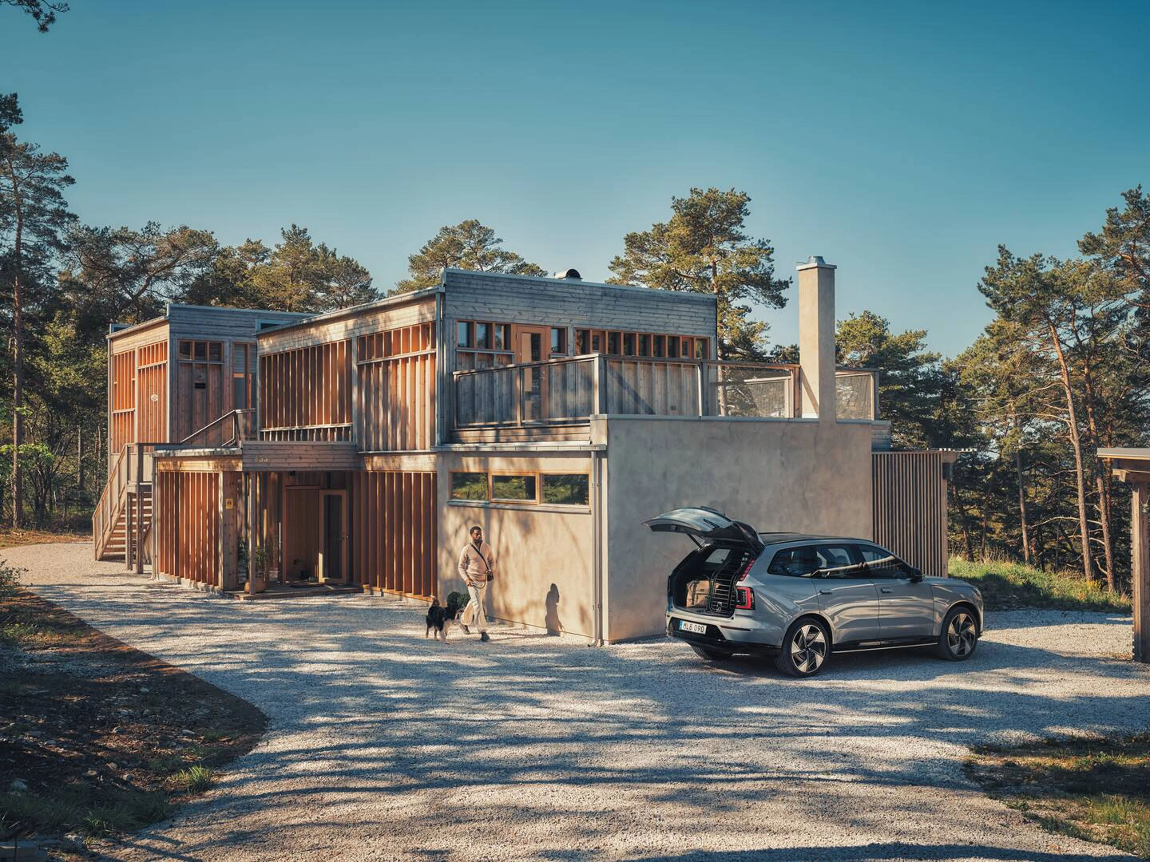 En stor bil fra Volvo ses skråt bagfra fra siden med bagageklappen åben, ved siden af et designerhus i beige nuancer med træer i baggrunden.