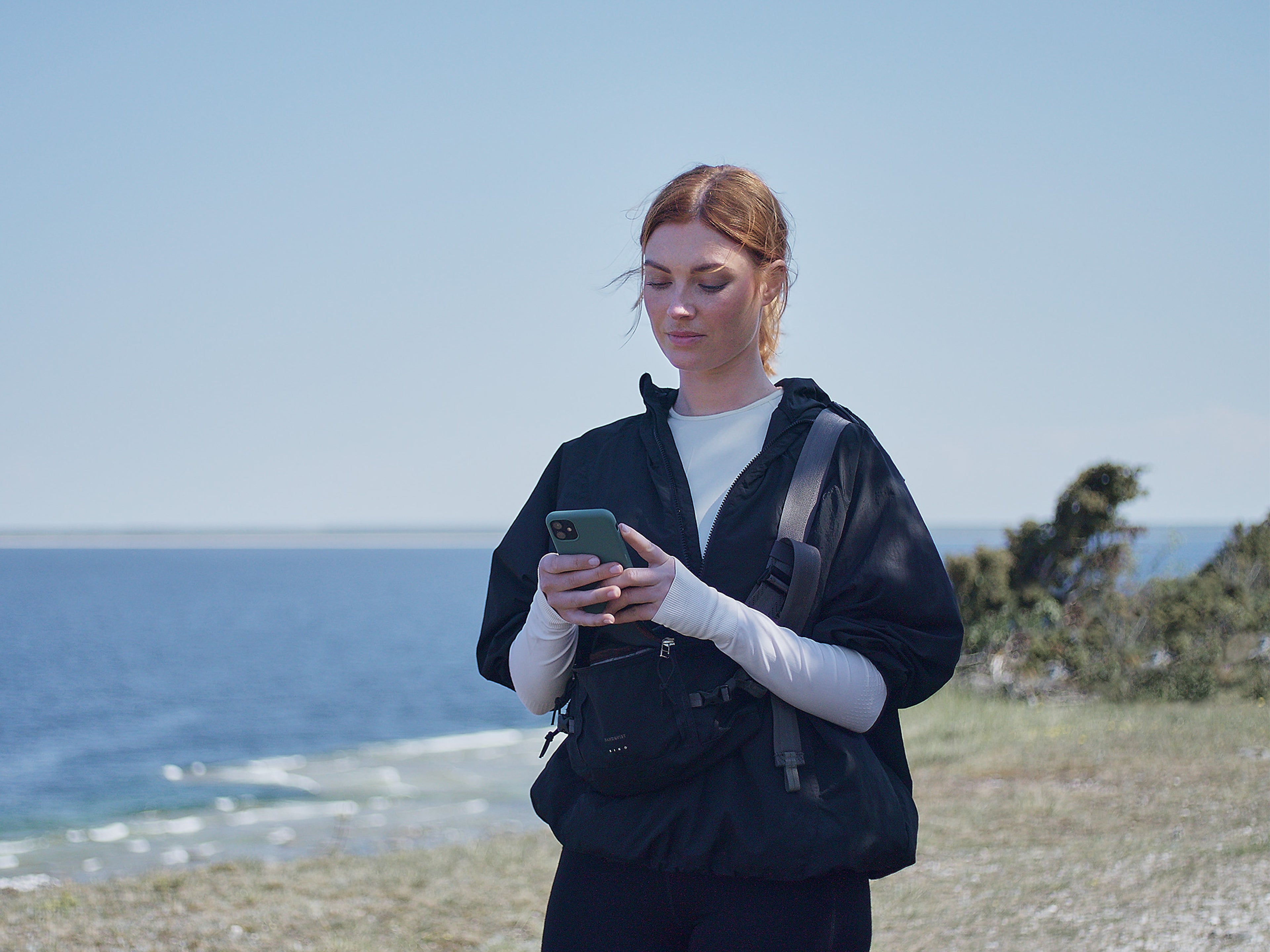 Punertavatukkainen nainen seisoo rannalla ja katsoo kädessään olevaa puhelinta.