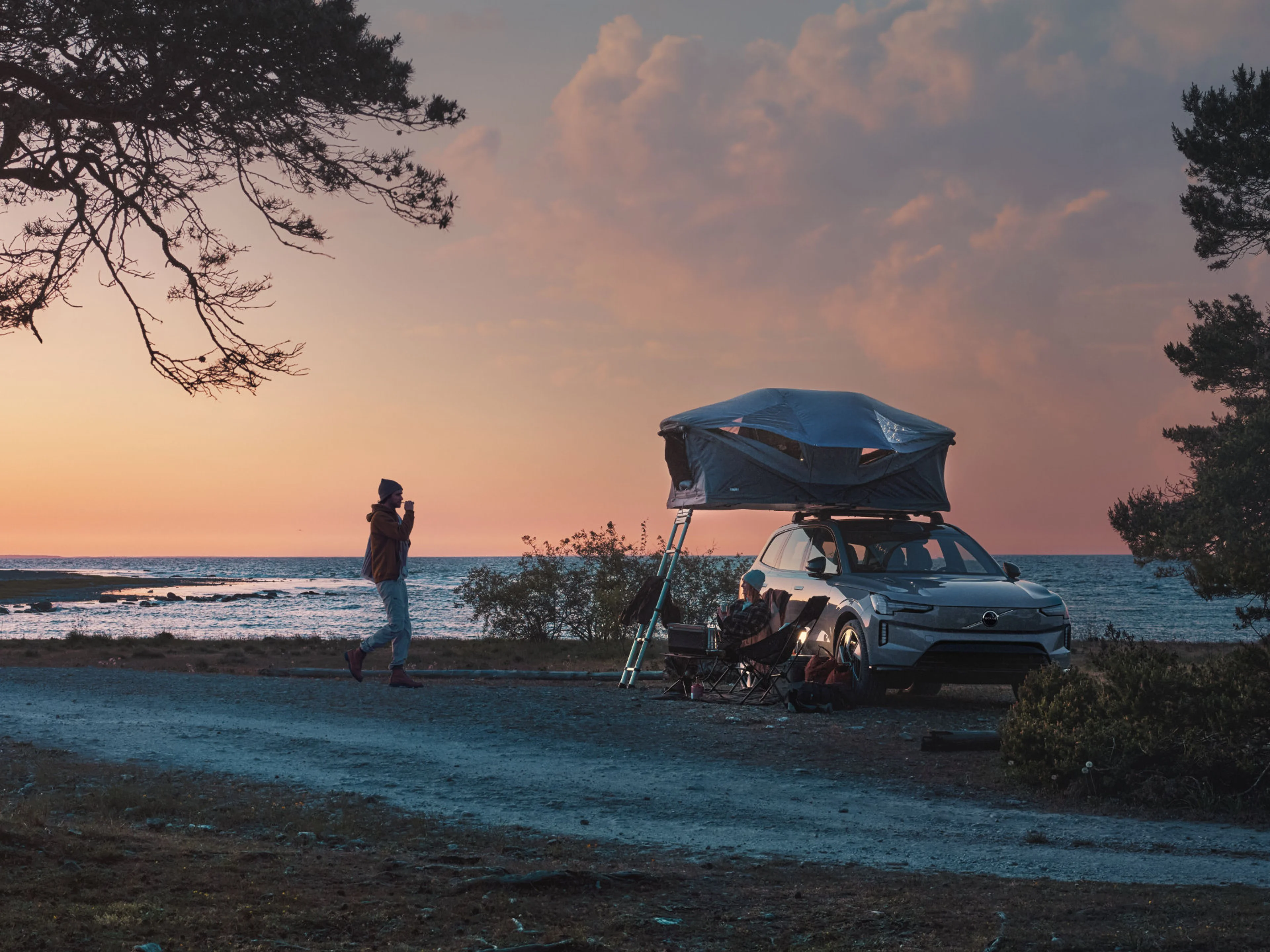 Ein Volvo EX90 mit ausgeklapptem Dachzelt steht im Abendlicht an einer mediterranen Küste zwischen Zedern. Zwei Personen genießen die Abendstimmung.