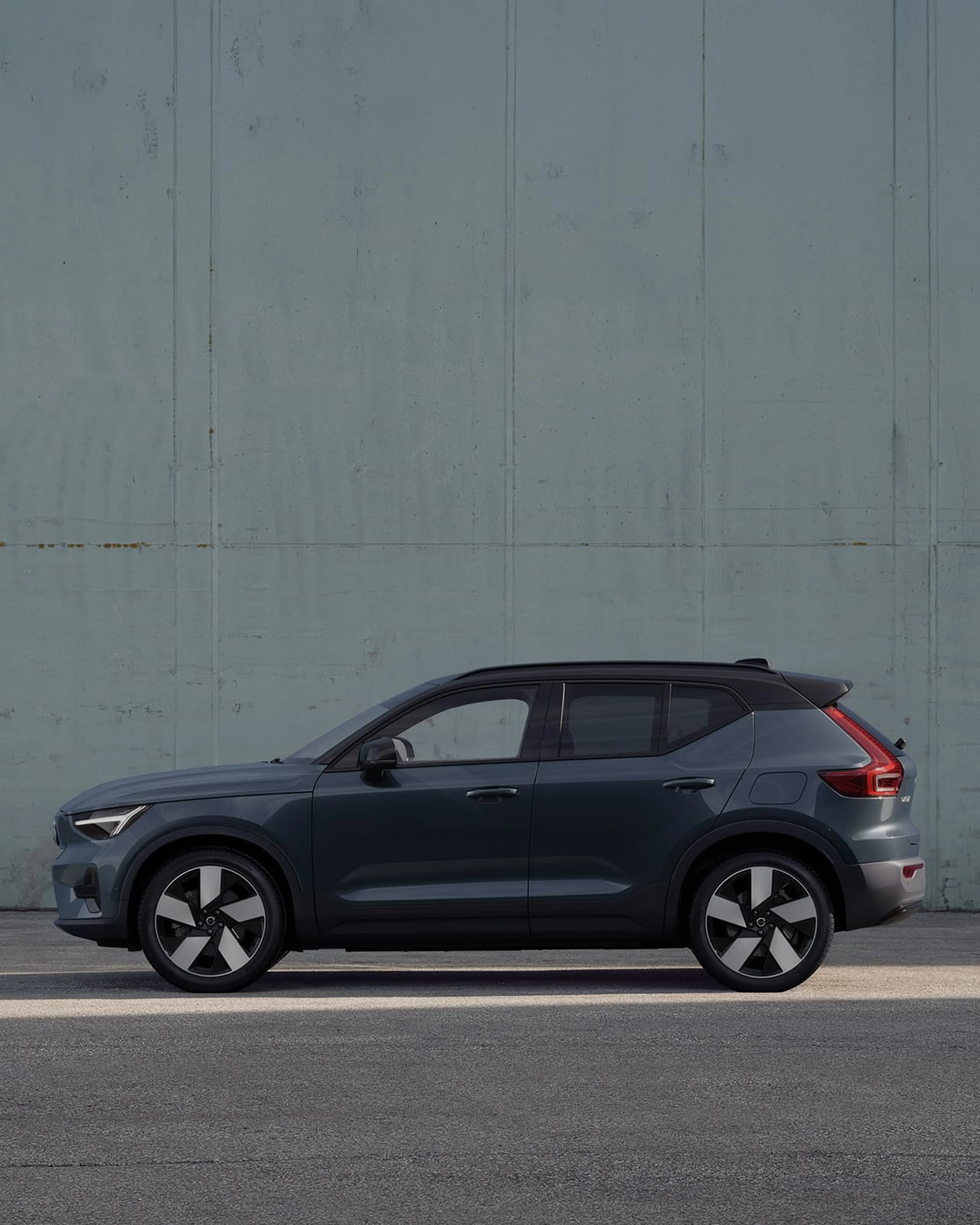 Vue de profil d'une Volvo EX40 en couleur Denim Blue avec toit noir garée devant un mur en béton gris clair.