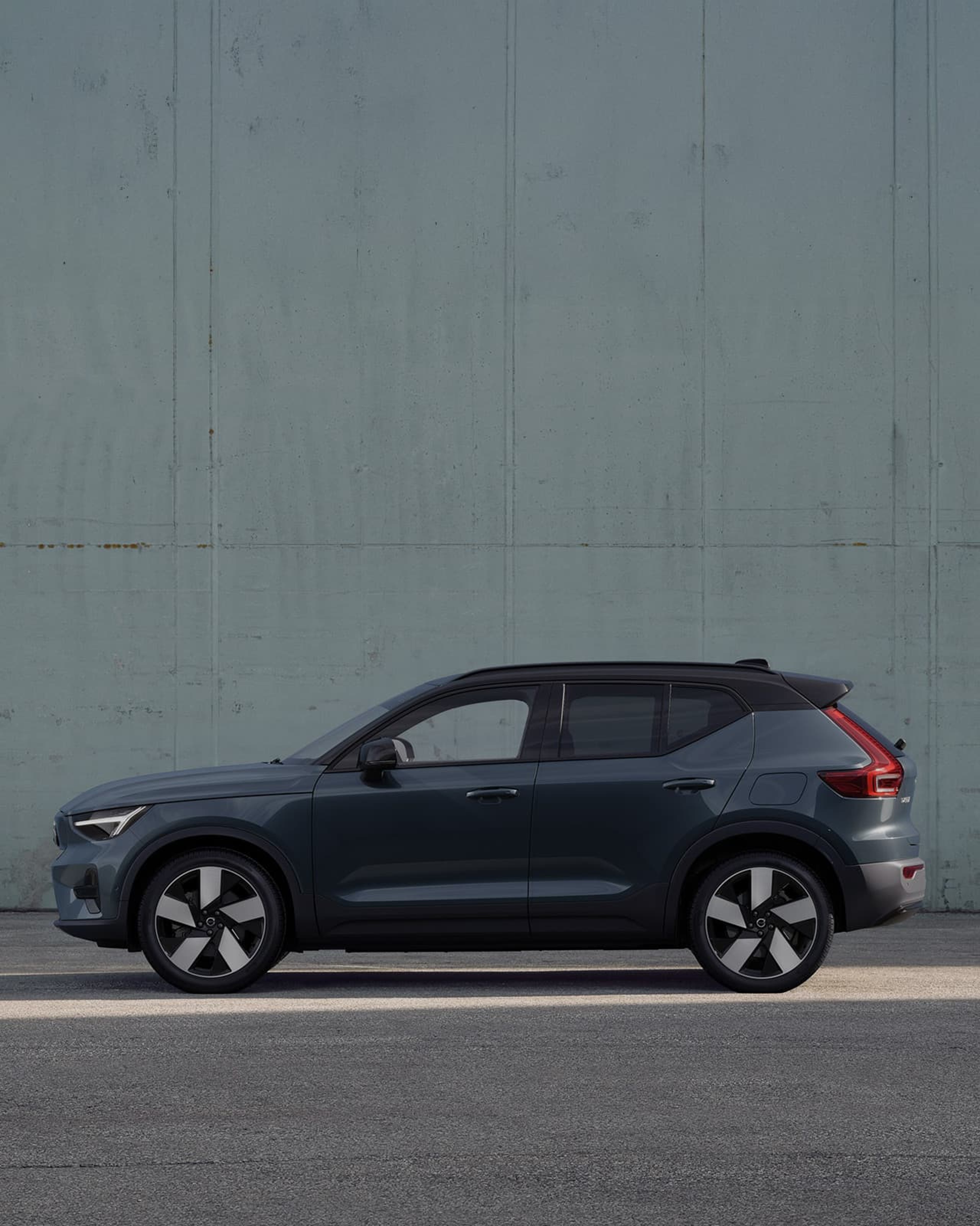 Zijaanzicht van een Volvo EX40 in de kleur Denim Blue met zwart dak geparkeerd voor een lichtgrijze betonnen muur.