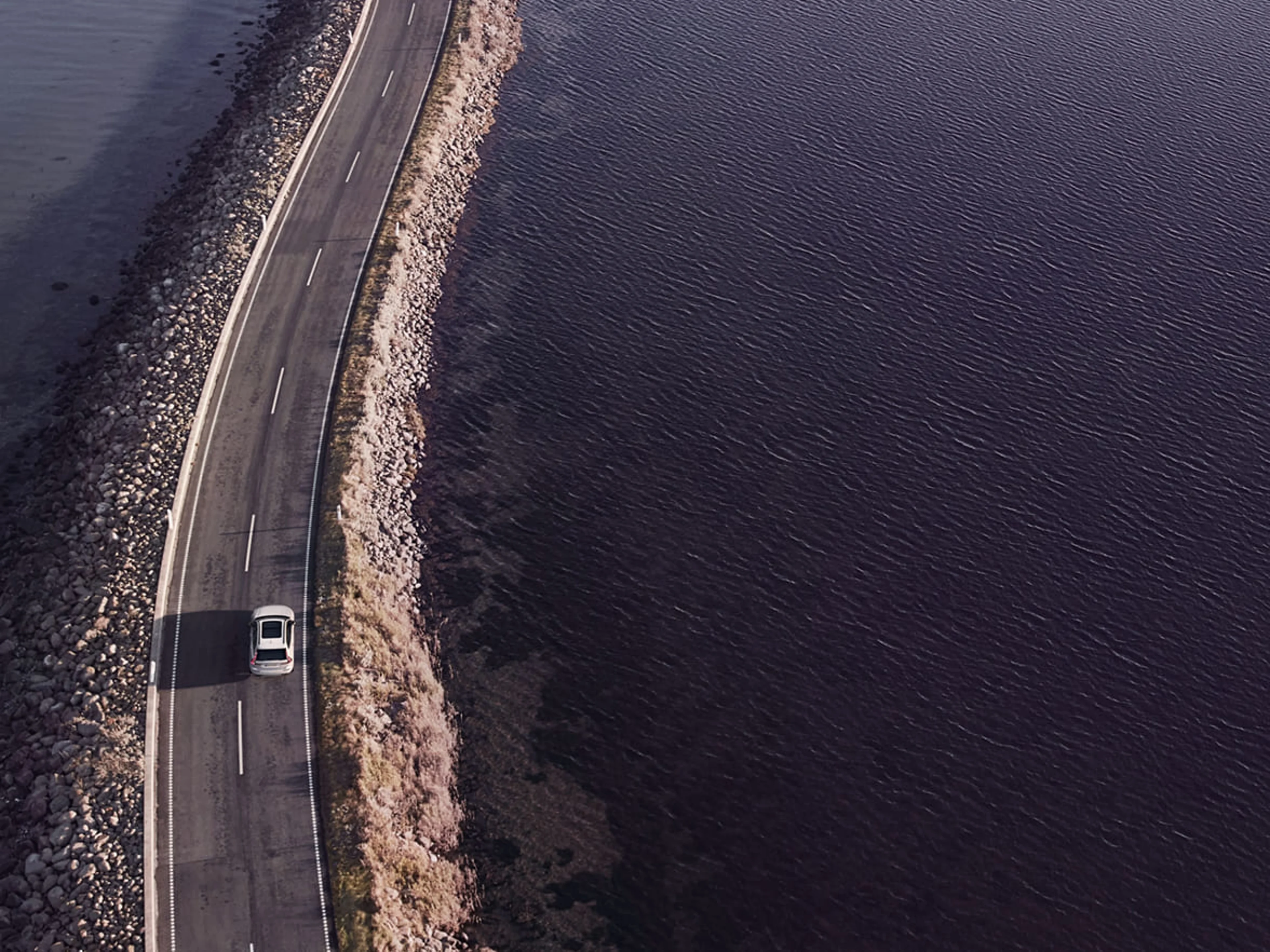 En bil kör på en smal landsväg som går längs en kustlinje med vatten på båda sidor. Vägen är kantad av stenar och vegetation, och bilden är tagen från ovan i ett fågelperspektiv.