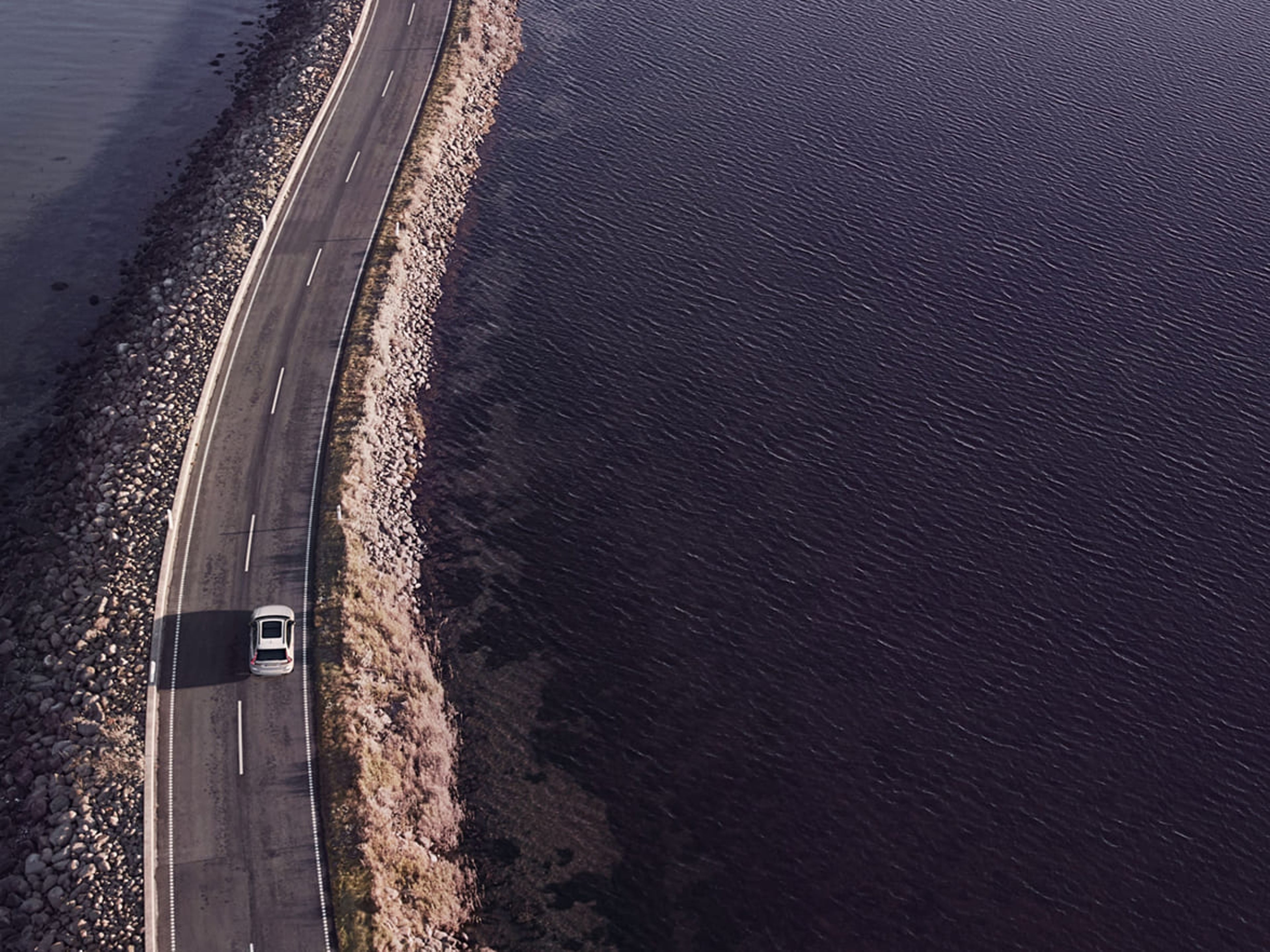 En bil kör på en smal landsväg som går längs en kustlinje med vatten på båda sidor. Vägen är kantad av stenar och vegetation, och bilden är tagen från ovan i ett fågelperspektiv.