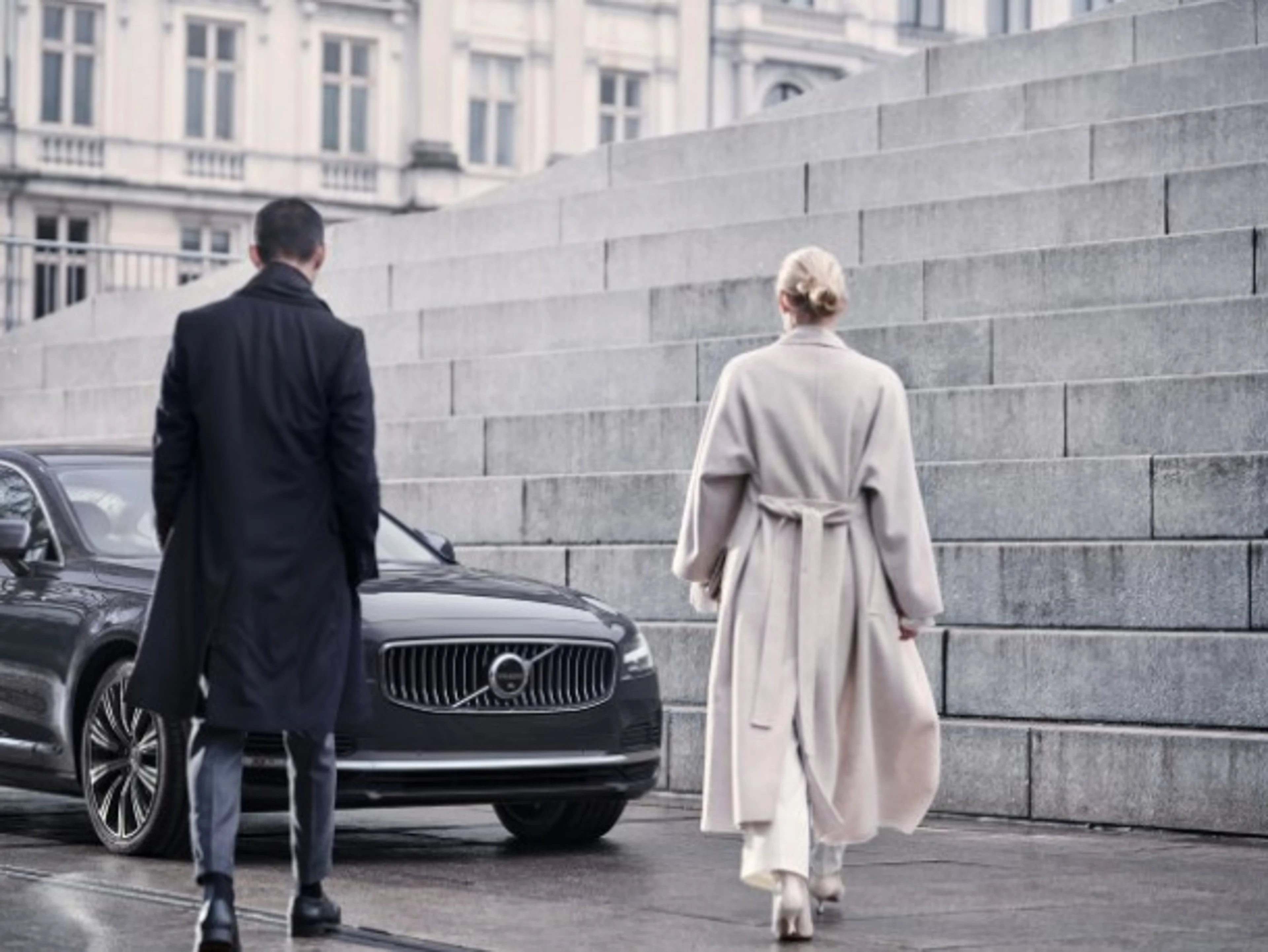 Elegantno oblečen moški in ženska hodita proti črnemu vozilu Volvo, parkiranemu ob modernih kamnitih stopnicah v mestnem okolju.