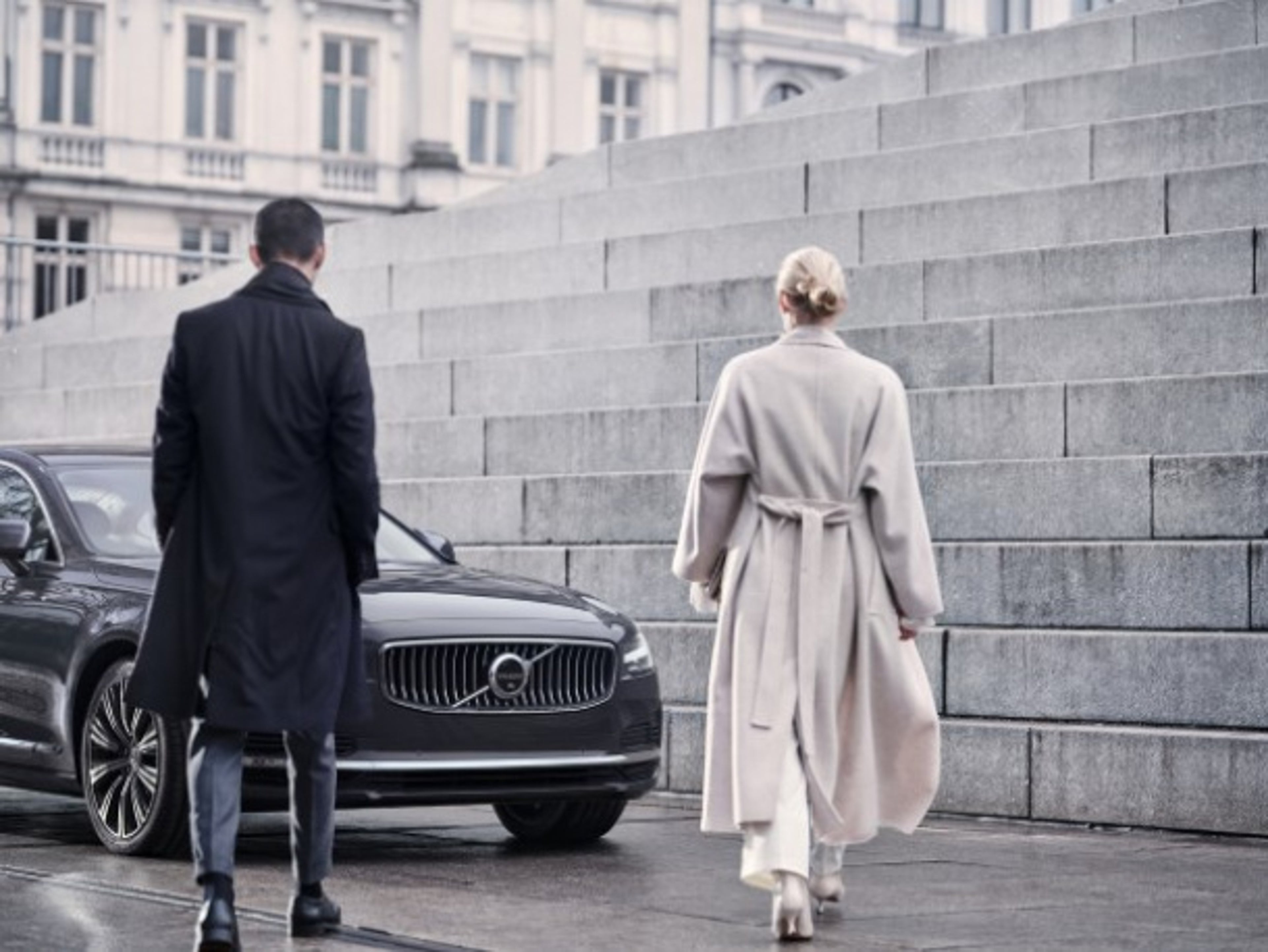 A man and a woman walking toward a dark Volvo vehicle parked next to stone steps in an urban setting.