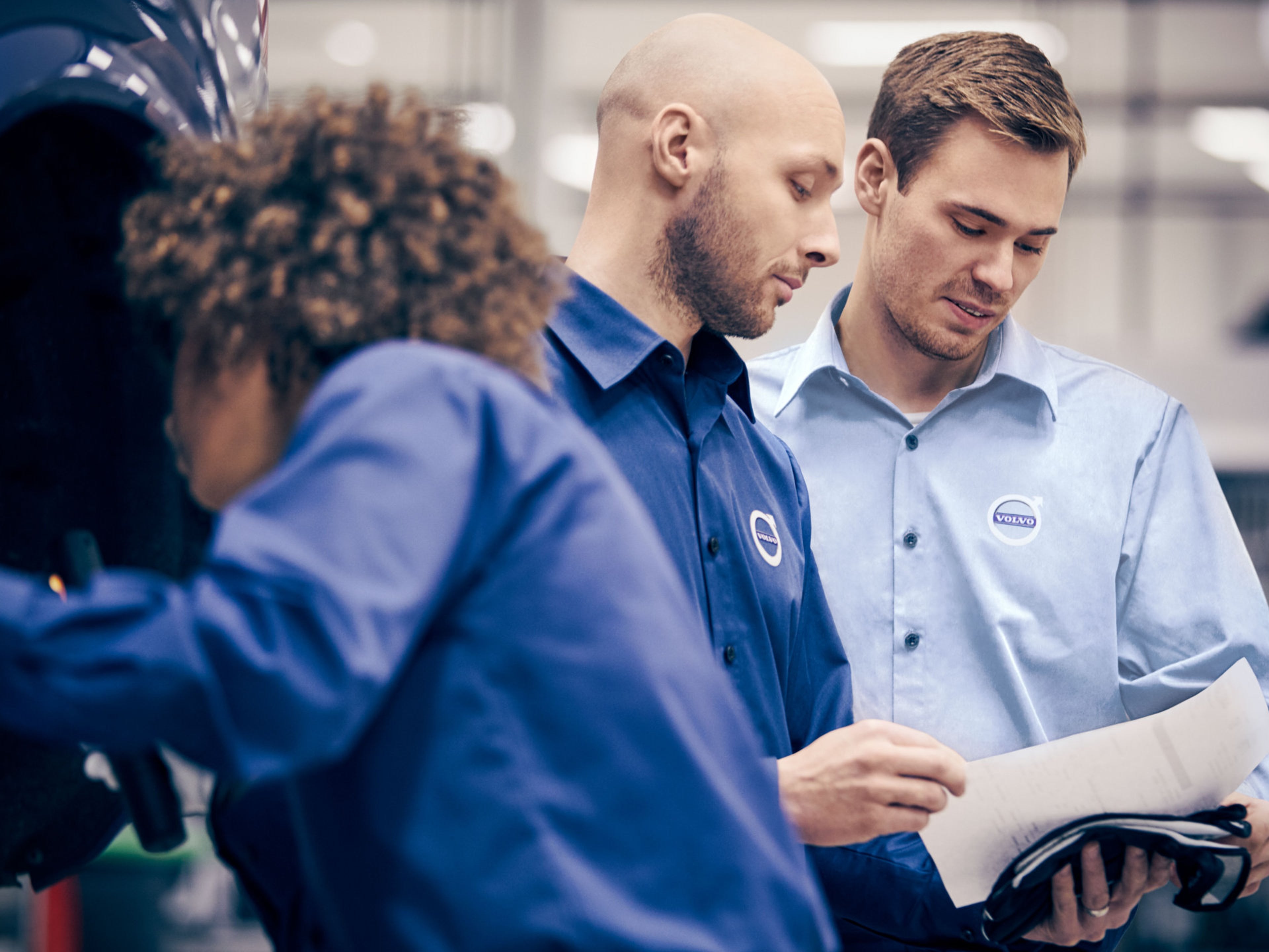 Zwei Volvo Mitarbeiter schauen zusammen auf einen Zettel, im Vordergrund eine Volvo Mitarbeiterin, welche gerade arbeitet.