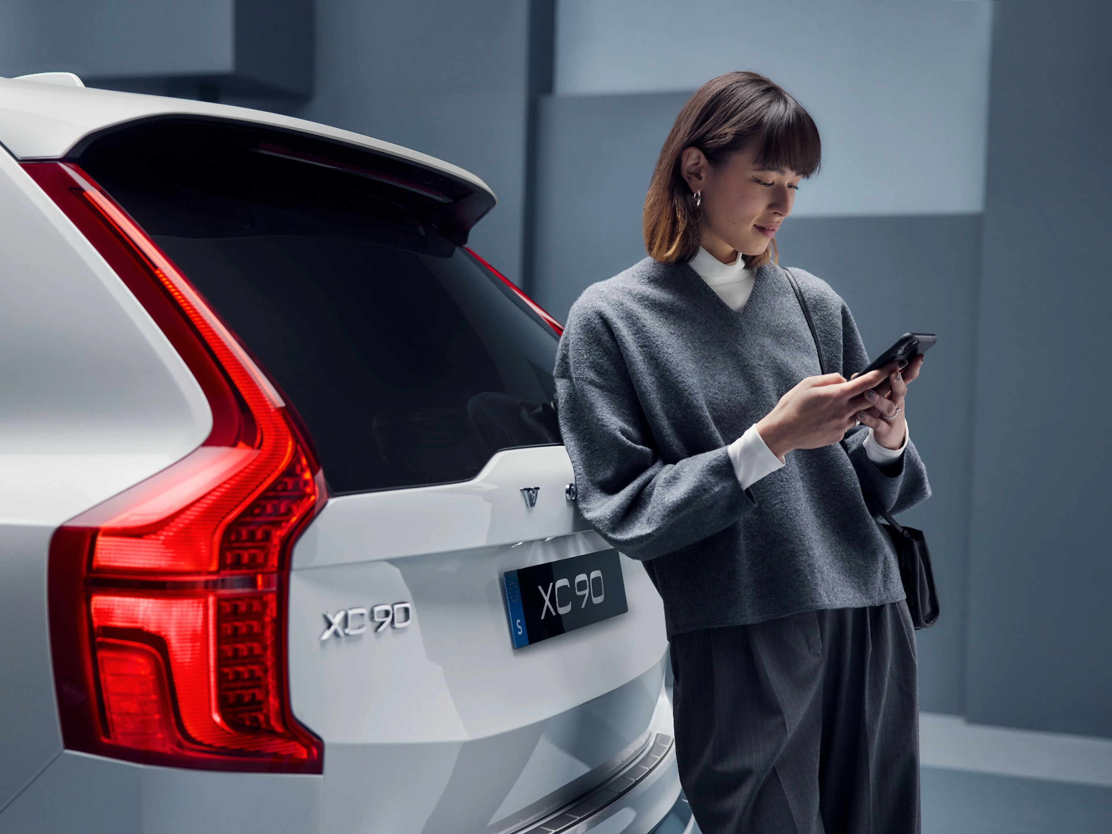 Eine Frau steht mit ihrem Smartphone in der Hand vor einem XC90 in weiß.