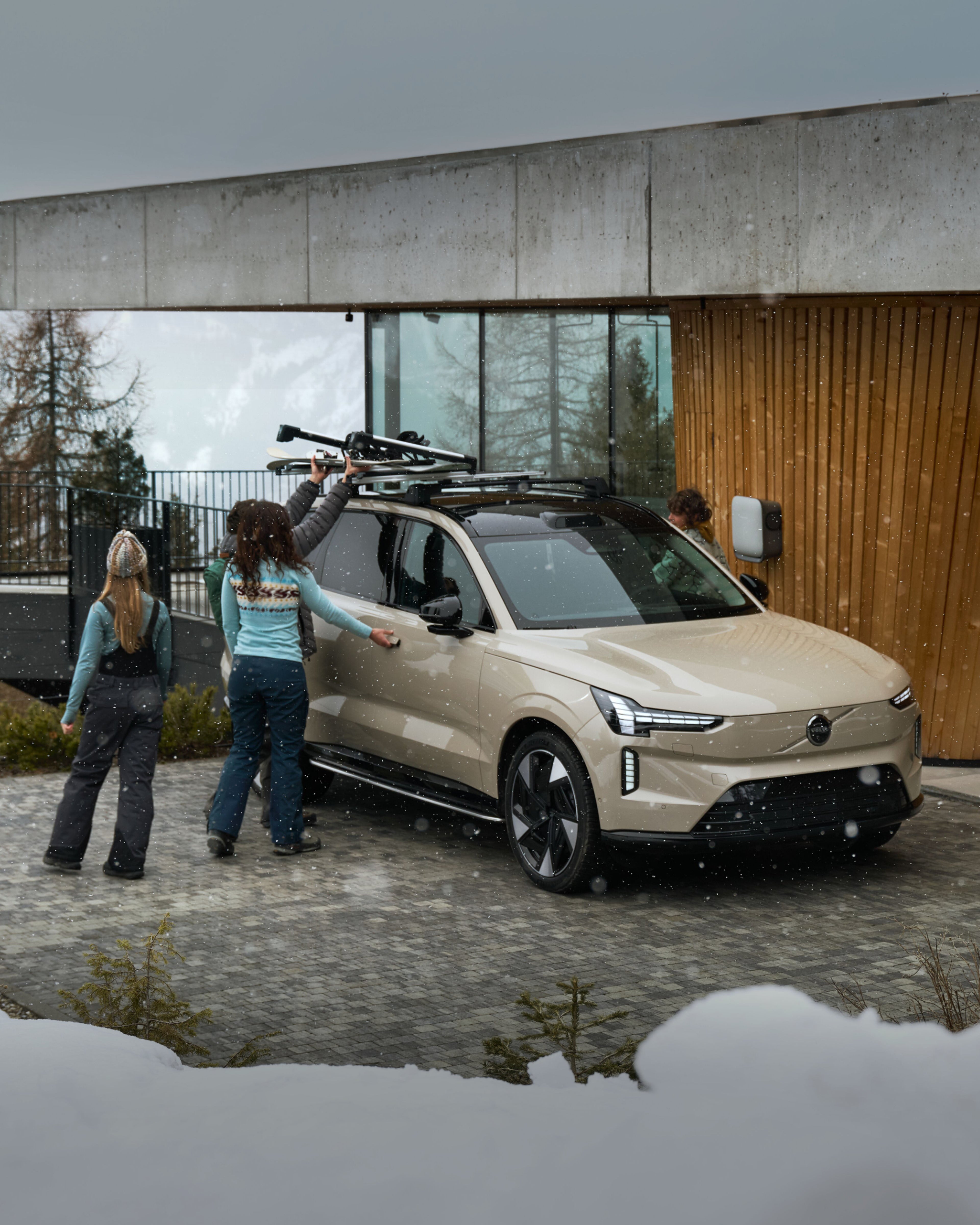 Zwei Personen und ein Mann stehen an einem beigen Volvo EX90 und bringen Skier auf dem Dach an.