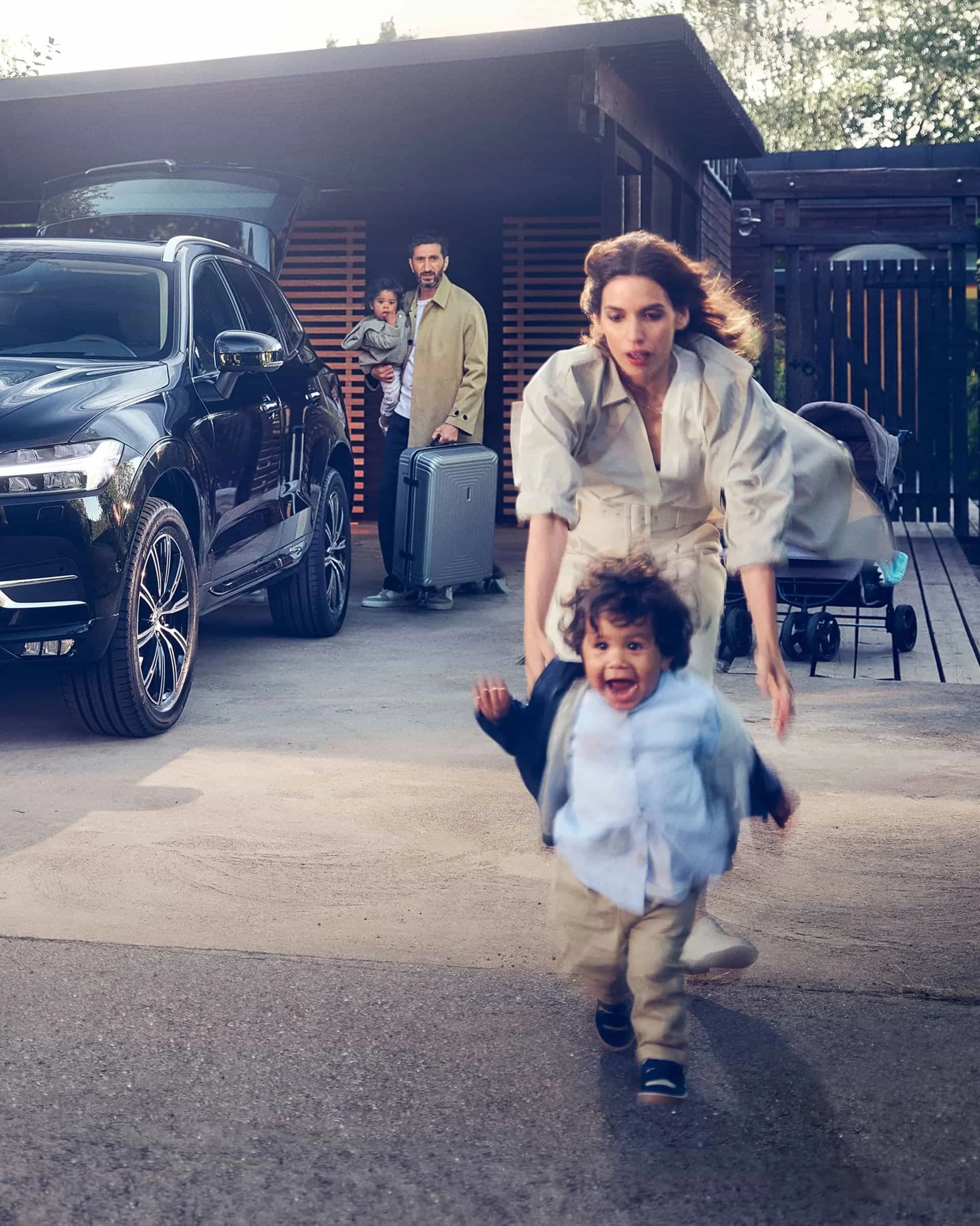 Une famille charge des bagages dans une Volvo noire devant une maison moderne, un enfant court avec enthousiasme devant.