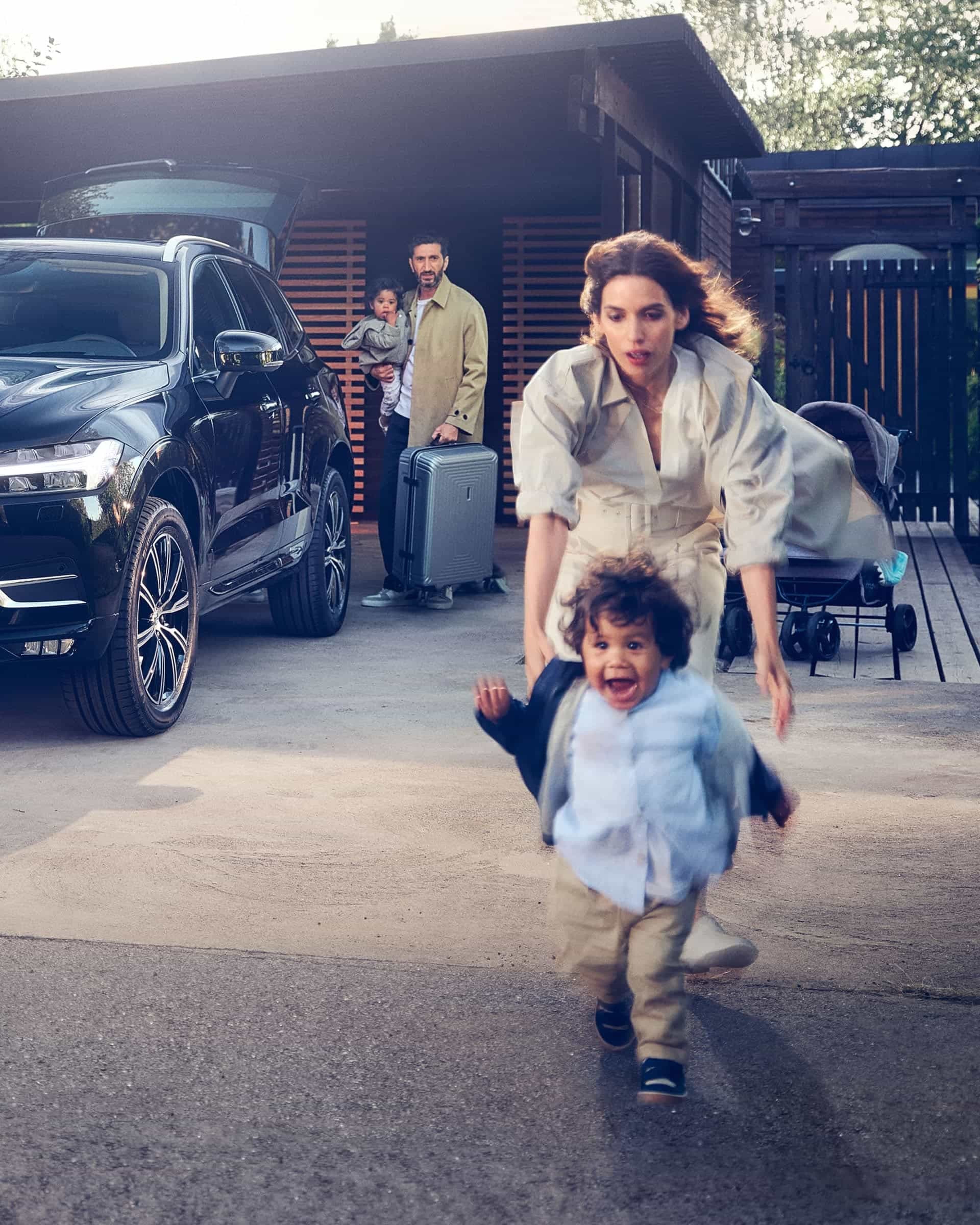 Une famille charge des bagages dans une Volvo noire devant une maison moderne, un enfant court avec enthousiasme devant.