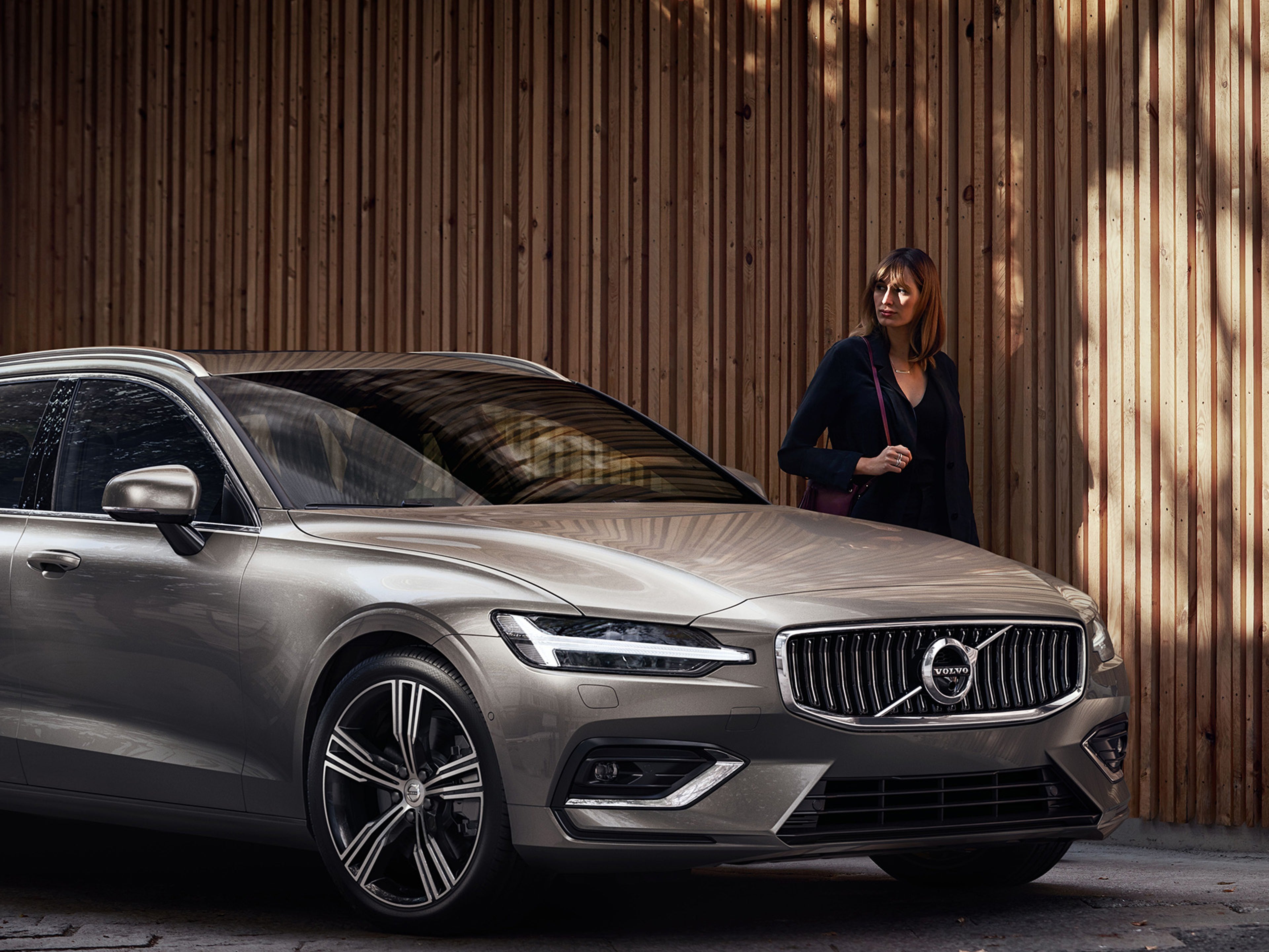 V60 cinzenta de frente, junto a uma parede de madeira, com uma mulher em segundo plano a olhar para o carro