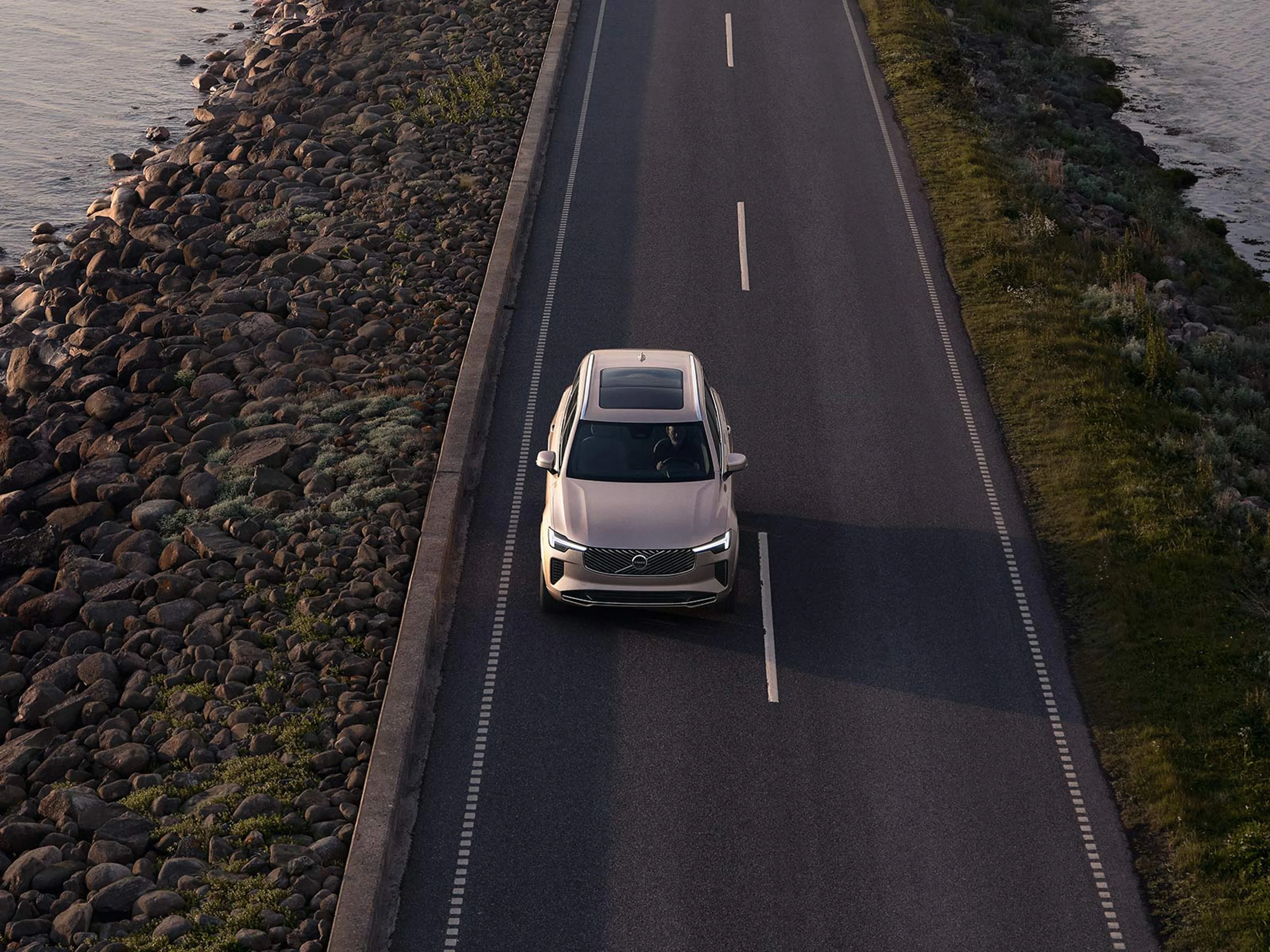 Vit Volvo XC90 sedd uppifrån på kustväg mellan vatten och klippor i kvällsljus.