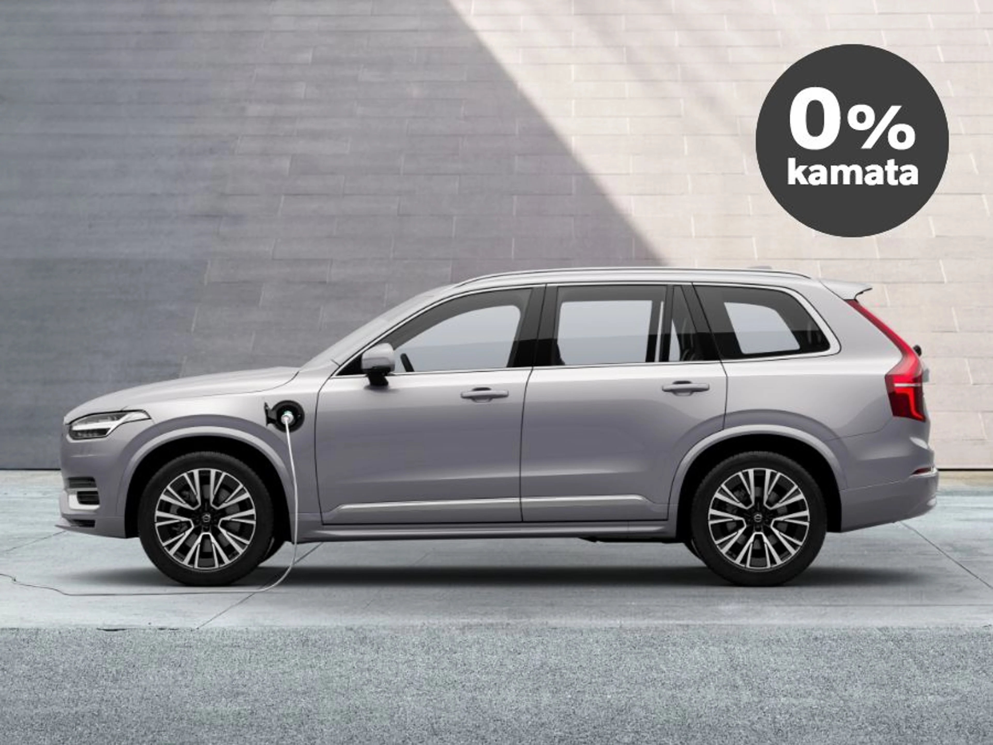 Srebrni Volvo XC90 plug-in hybrid priključen na punjenje ispred moderne zgrade, s oznakom '0% kamata' u crnom krugu u gornjem desnom kutu.
