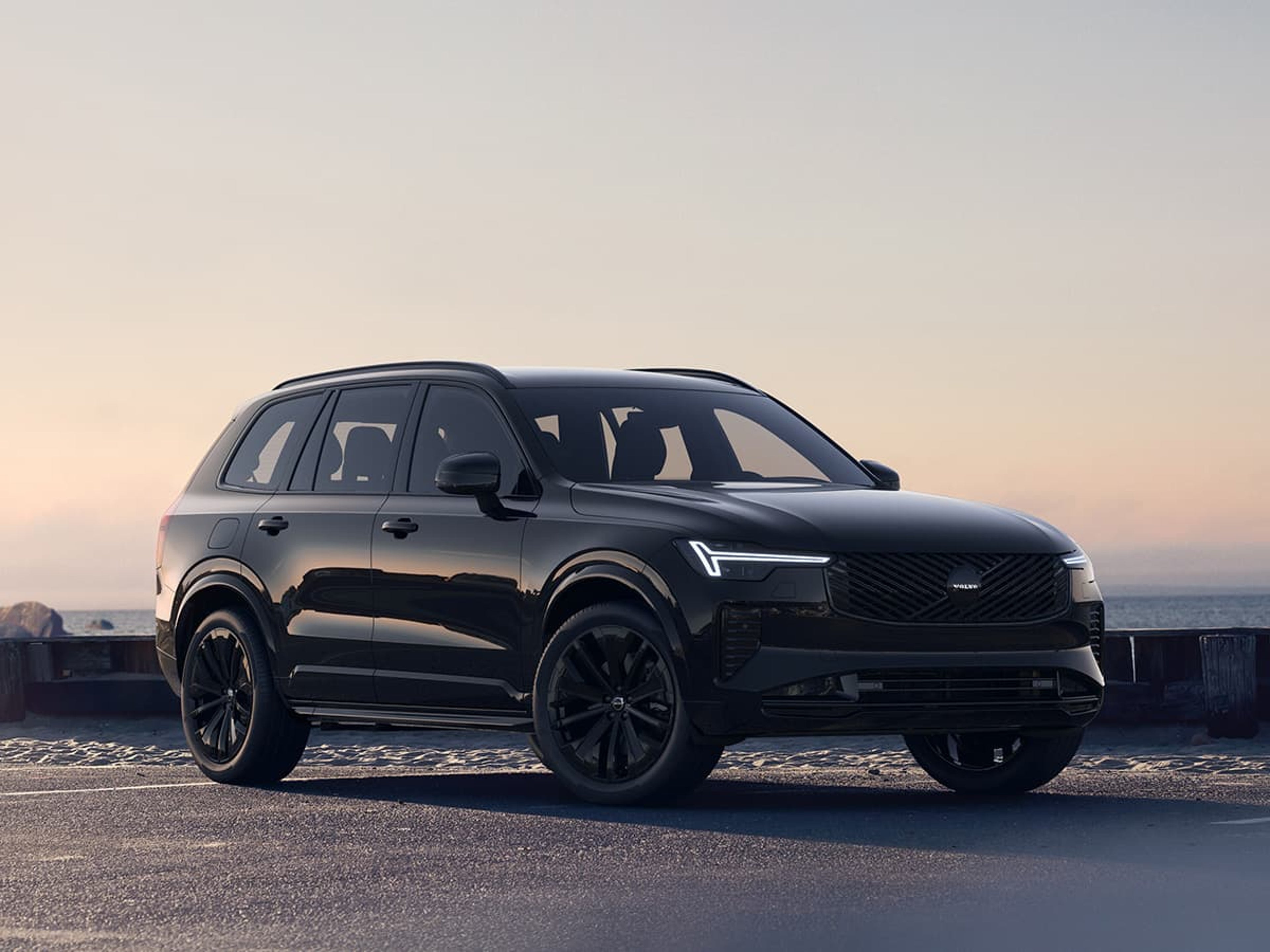 Vue de trois quarts avant d'un Volvo XC90 Black Edition entièrement noir, garé en bord de mer au coucher du soleil.