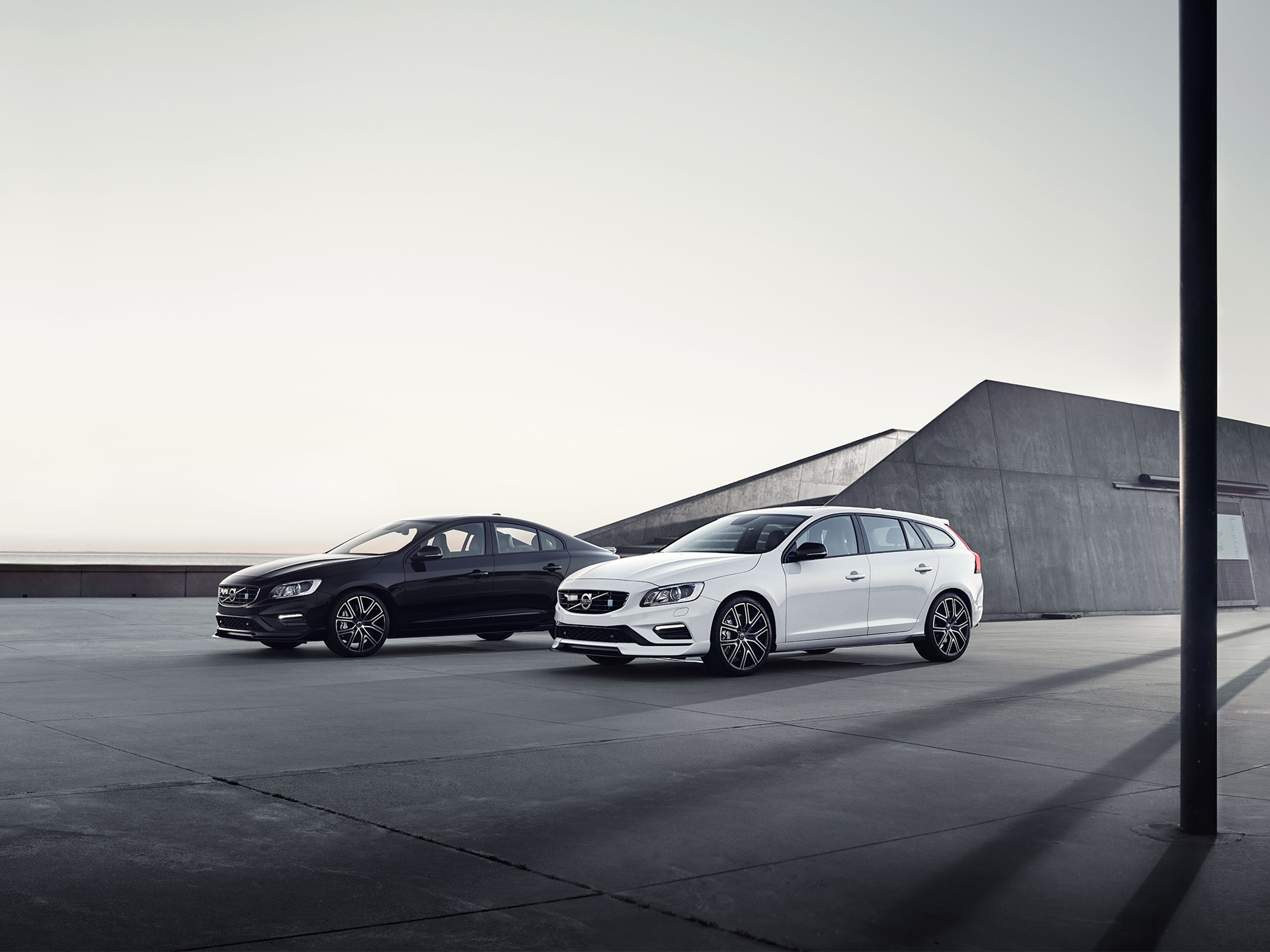 Musta Volvo S60 ja valkoinen Volvo V60 parkissa tasanteella, takana geometrinen betonirakennus.