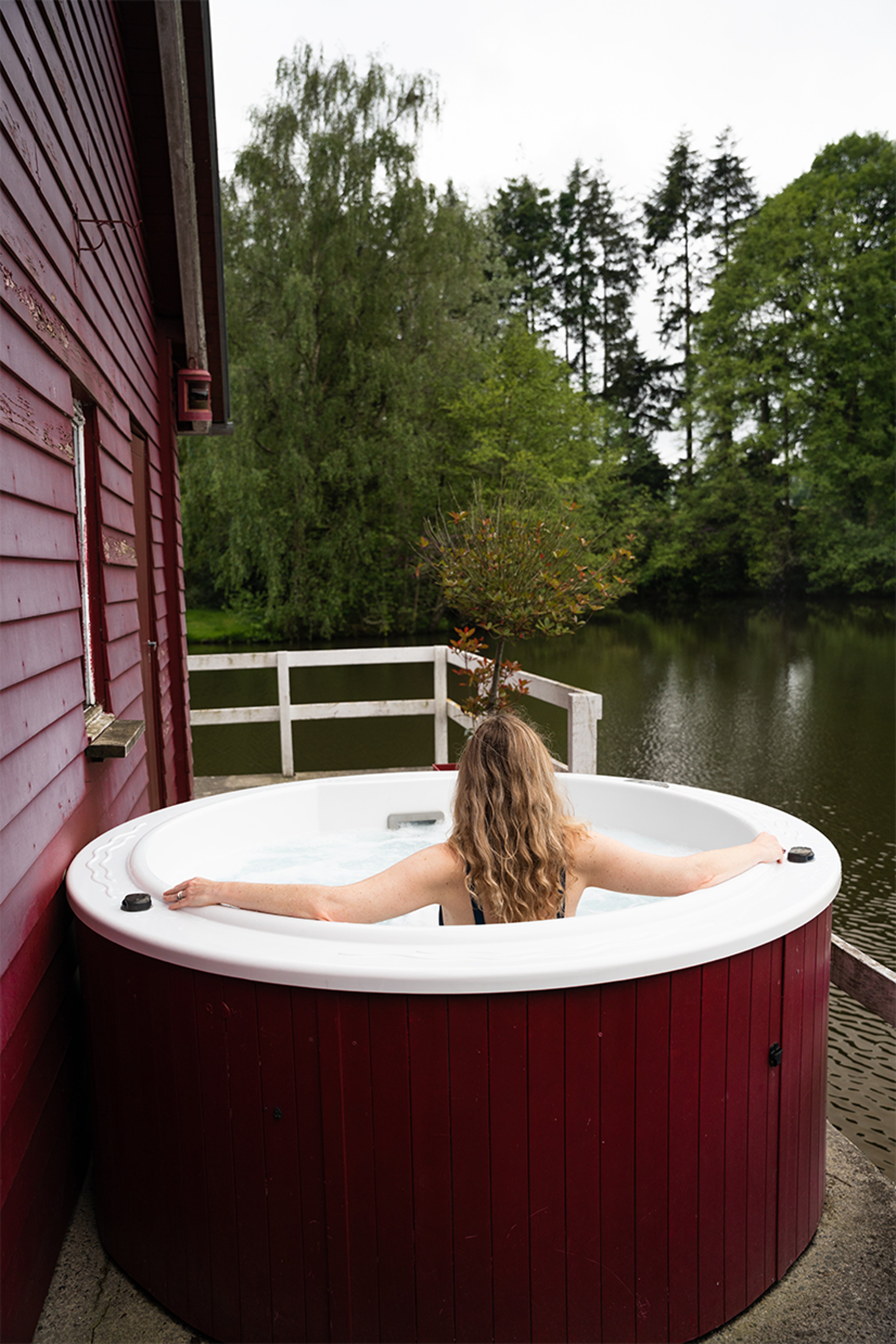 Personne dans jacuzzi rouge près d’une maison en bois, vue sur lac et forêt en arrière-plan.