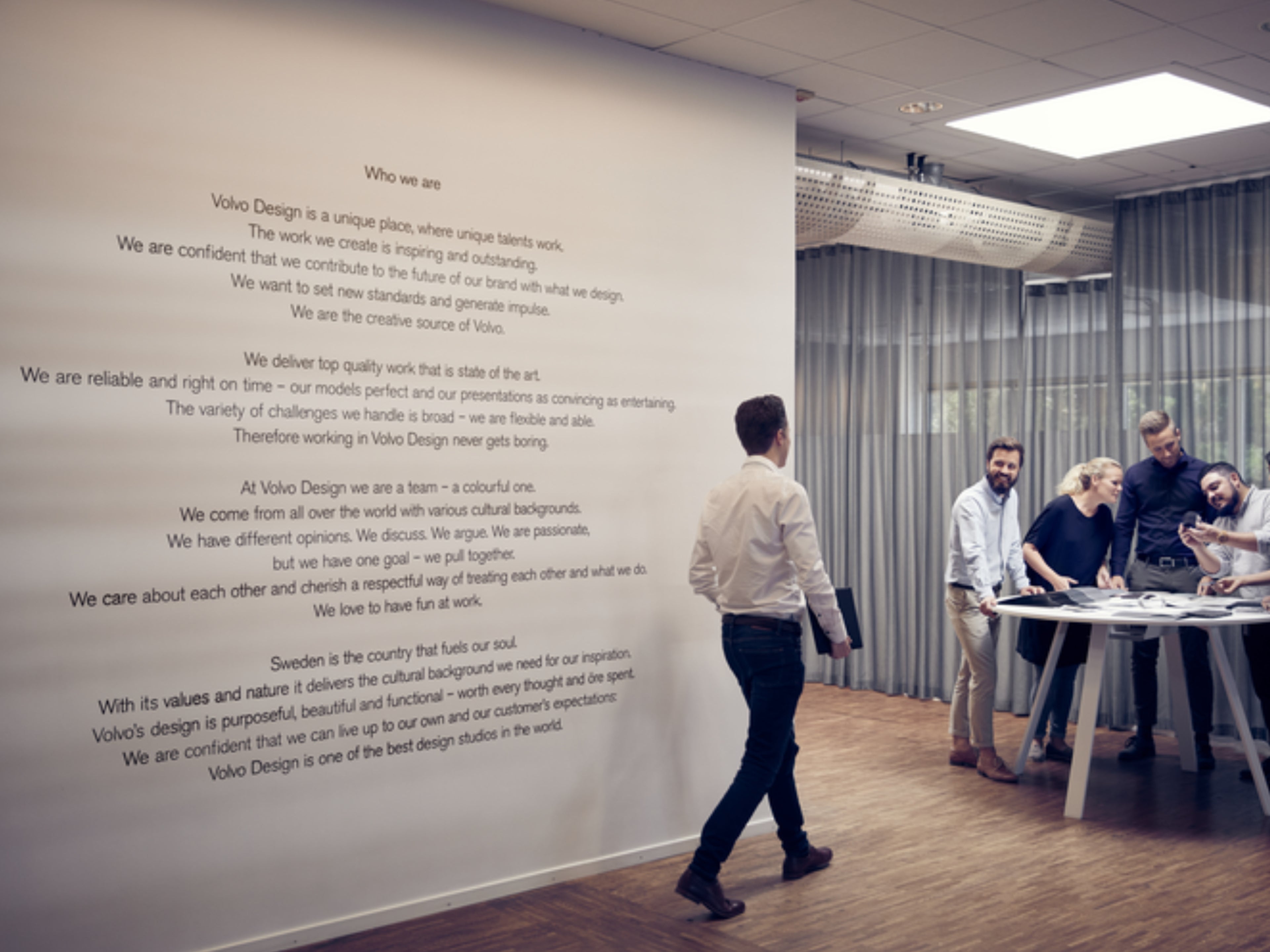 Vier Volvo Mitarbeiter stehen in einem Büro rund um einen Tisch, während ein weiterer, männlicher Mitarbeiter mit Laptop auf sie zuschreitet.