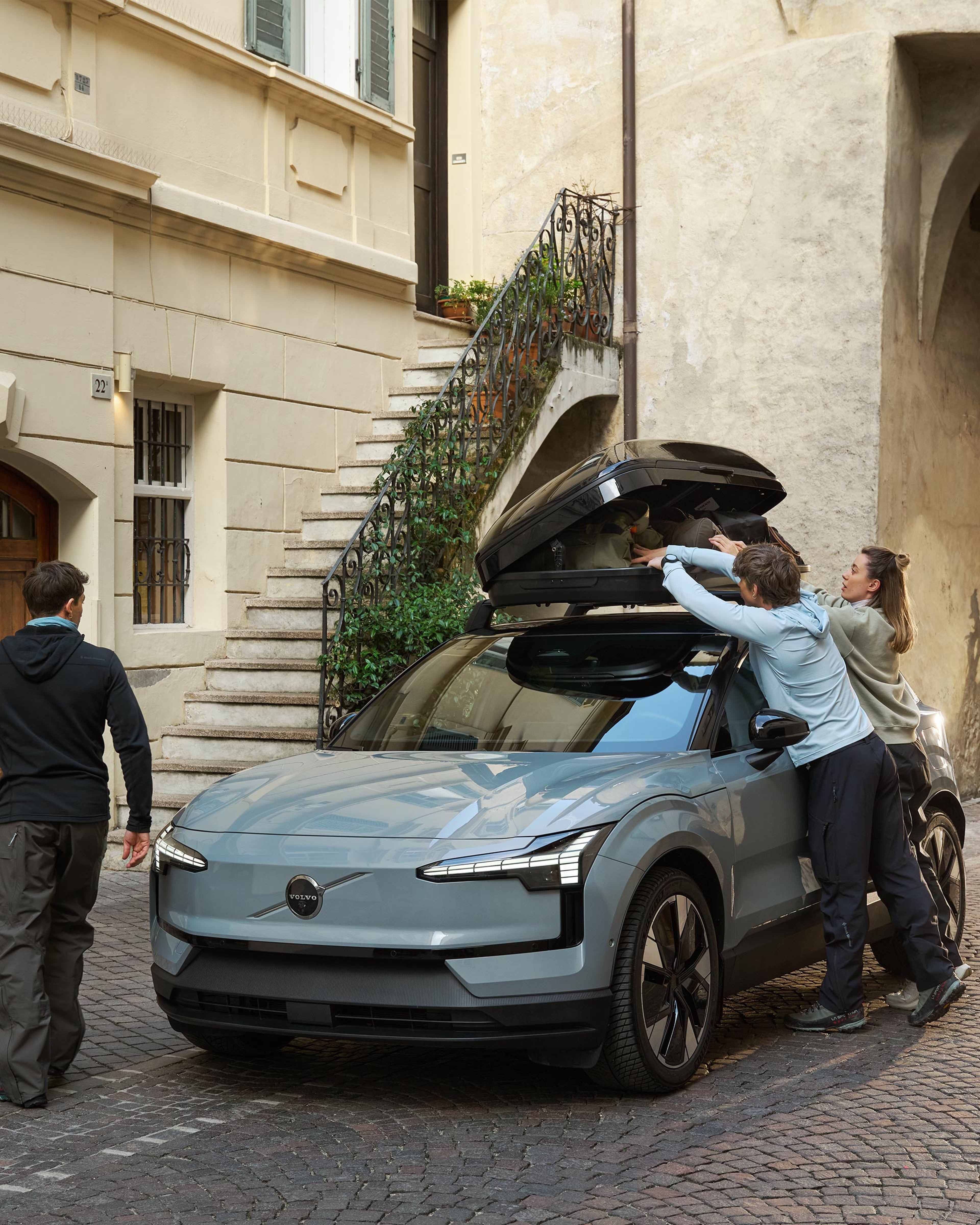 Nainen ja mies pakkaavat tavaroita Vapor grey -värisen Volvo EX30 täyssähköisen katumaasturin katolle kiinnitettyyn kattolaatikkoon Keski-Eurooppalaisen kivitalon pihalla. Toinen mies kiertää auton toiselle puolelle.