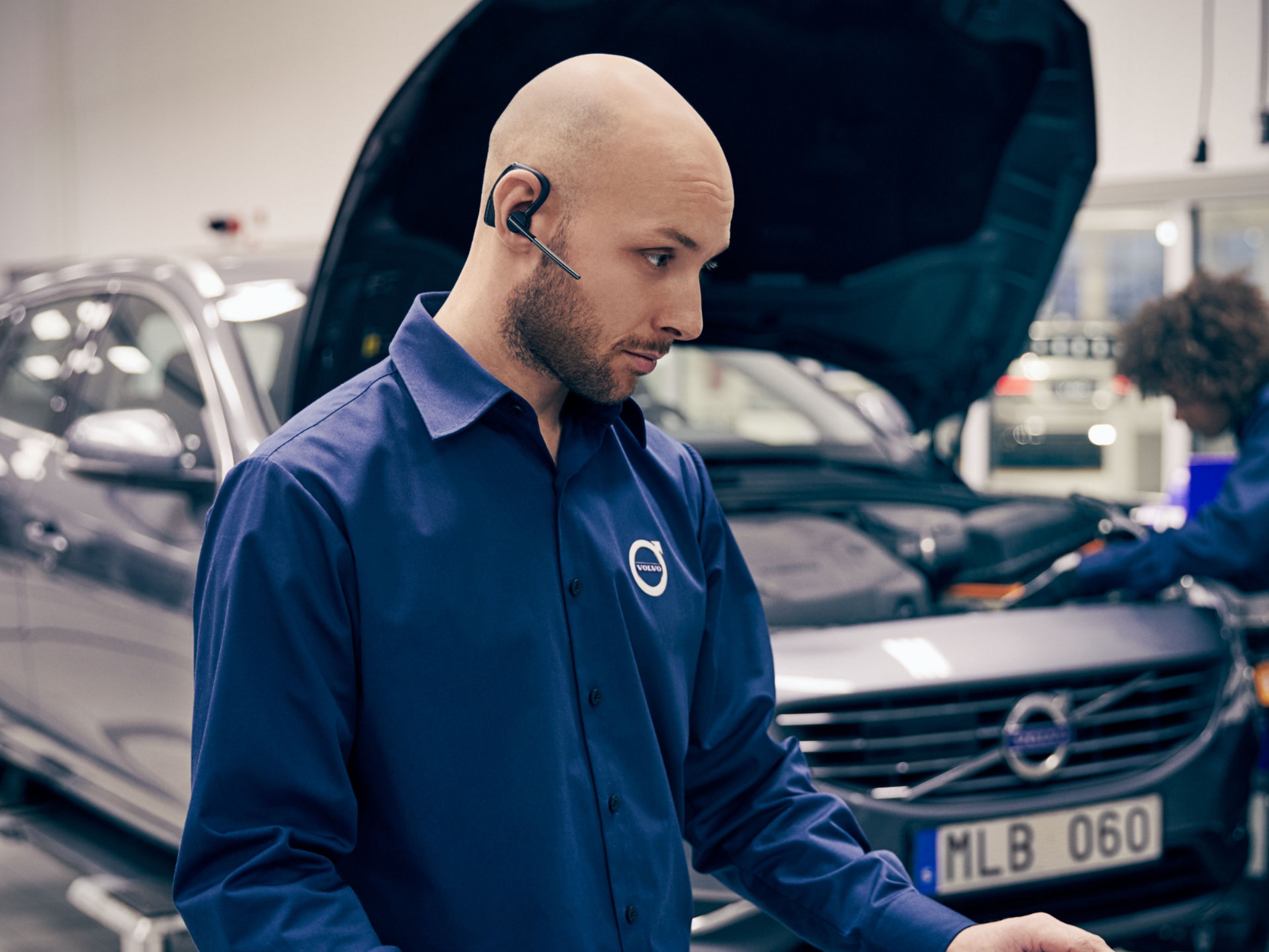 Ein männlicher Volvo Personal Service Techniker steht mit Telefon-Headset vor der geöffneten Haube eines Volvo innerhalb einer Werkstatt. Eine weibliche Mechanikerin arbeitet im Motorraum.