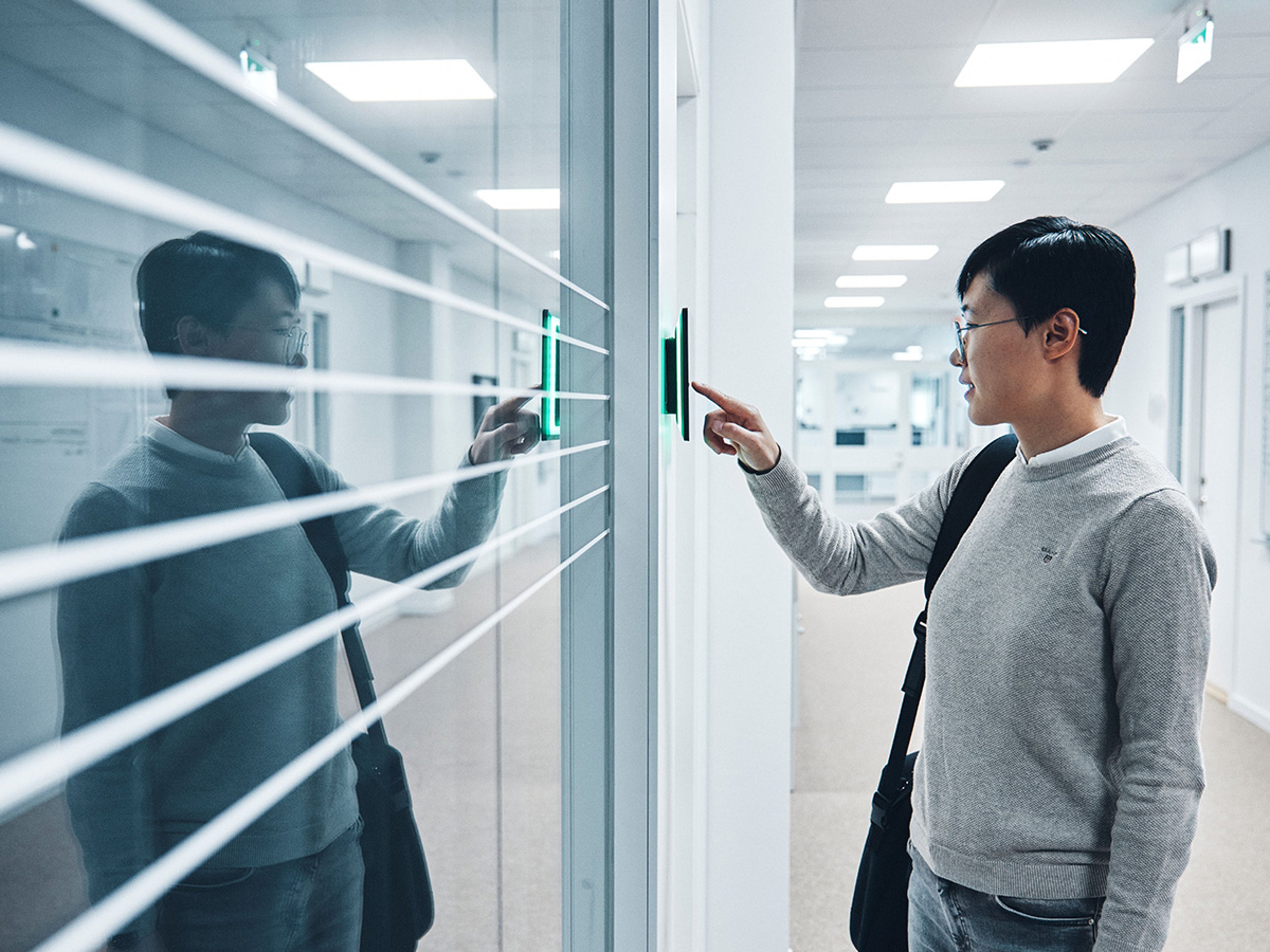 Een persoon met een grijze trui en een zwarte schoudertas gebruikt een digitaal toegangspaneel in een kantooromgeving.