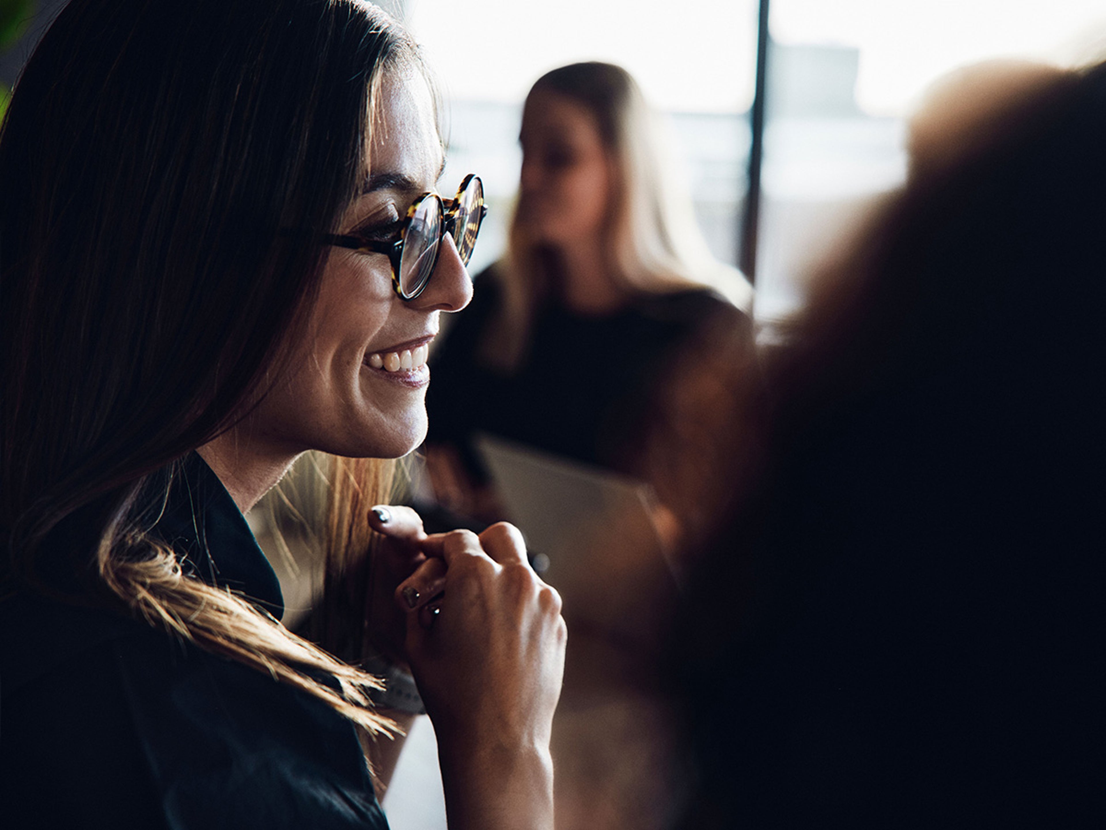 Een glimlachende vrouw met een bril en donker haar draait zich naar rechts, terwijl twee andere mensen op de achtergrond te zien zijn.