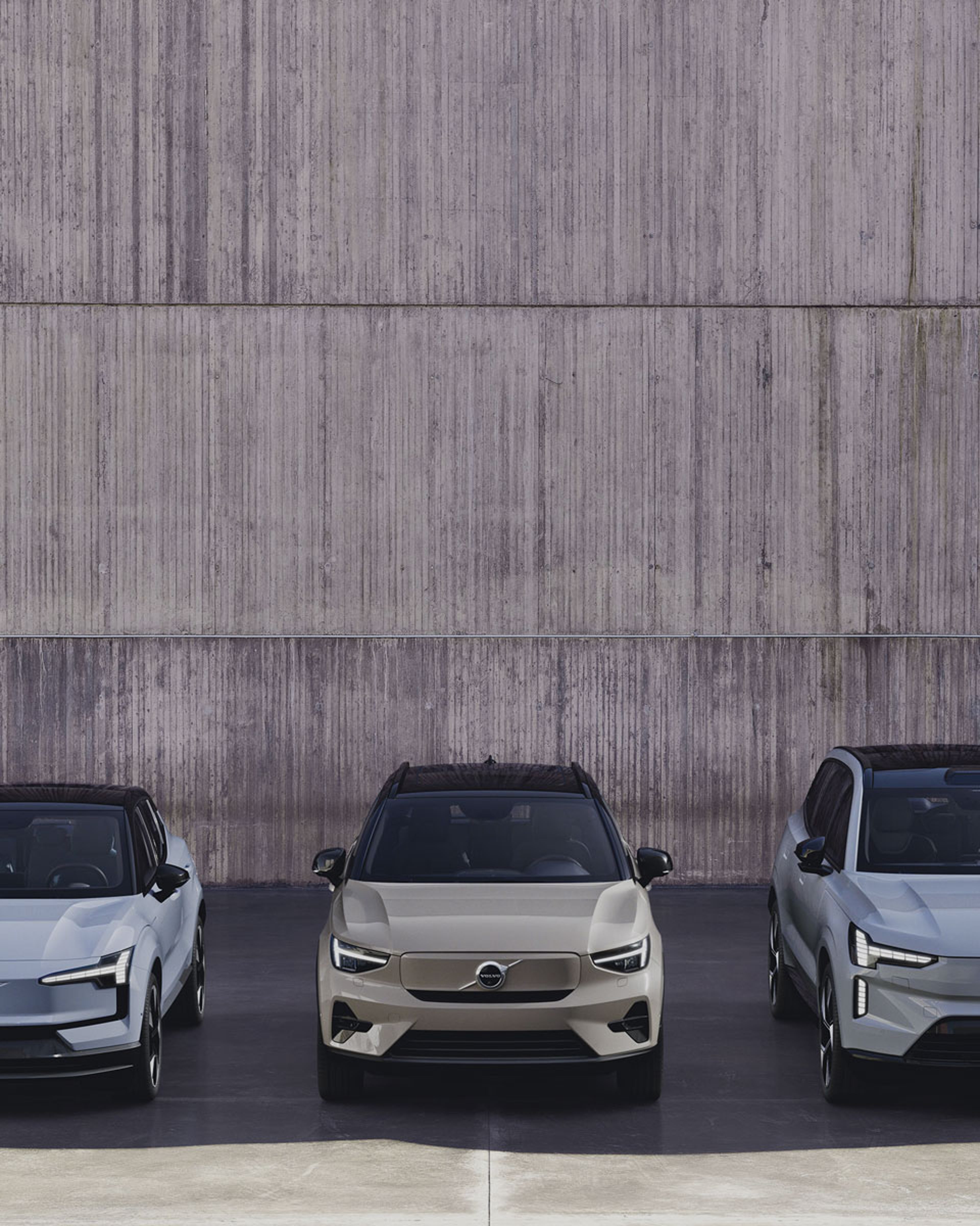 Een Volvo EX30, EX40 en een EX90 staan voor een betonnen muur geparkeerd.