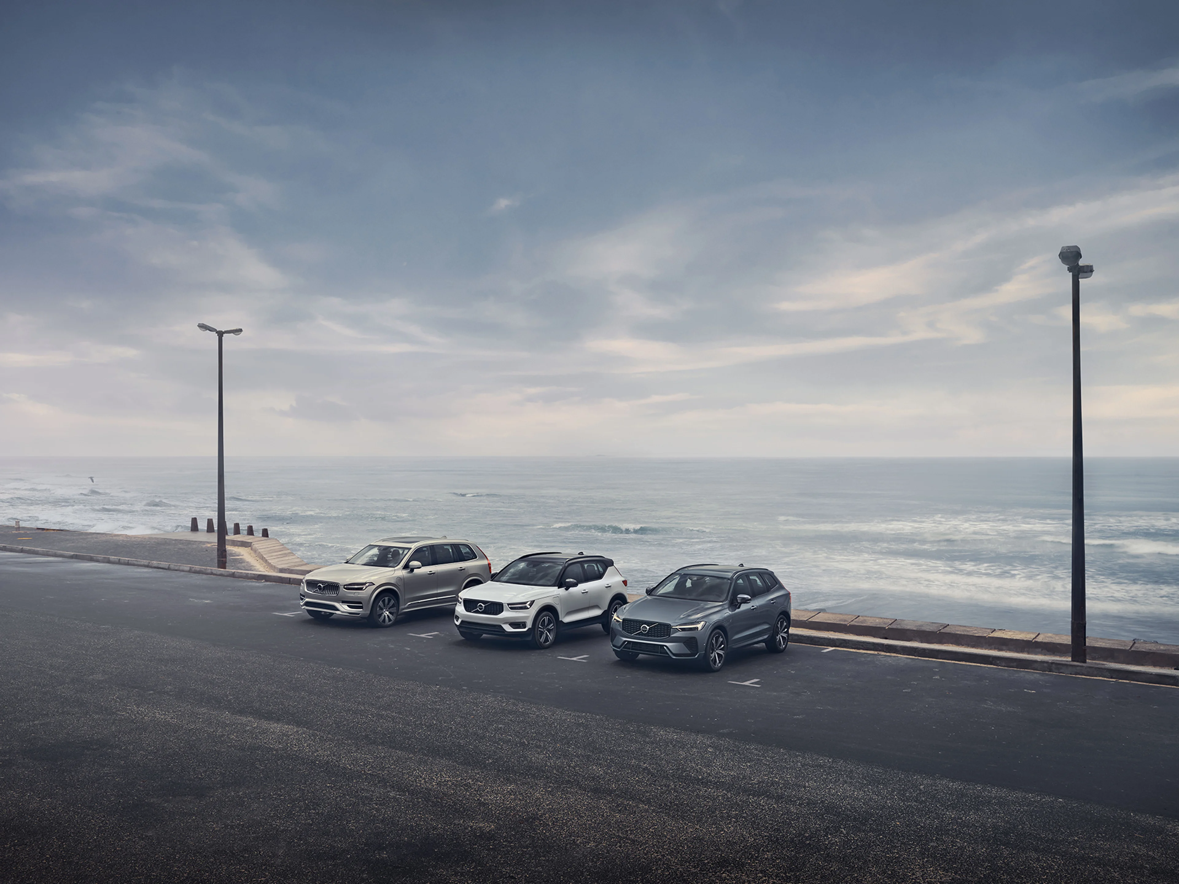 Ein champagnerfarbener Volvo EX90 steht rechts von einem weißen Volvo XC40. Neben diesem steht links ein blaugrauer Volvo XC60 vor einer Küstenkulisse.