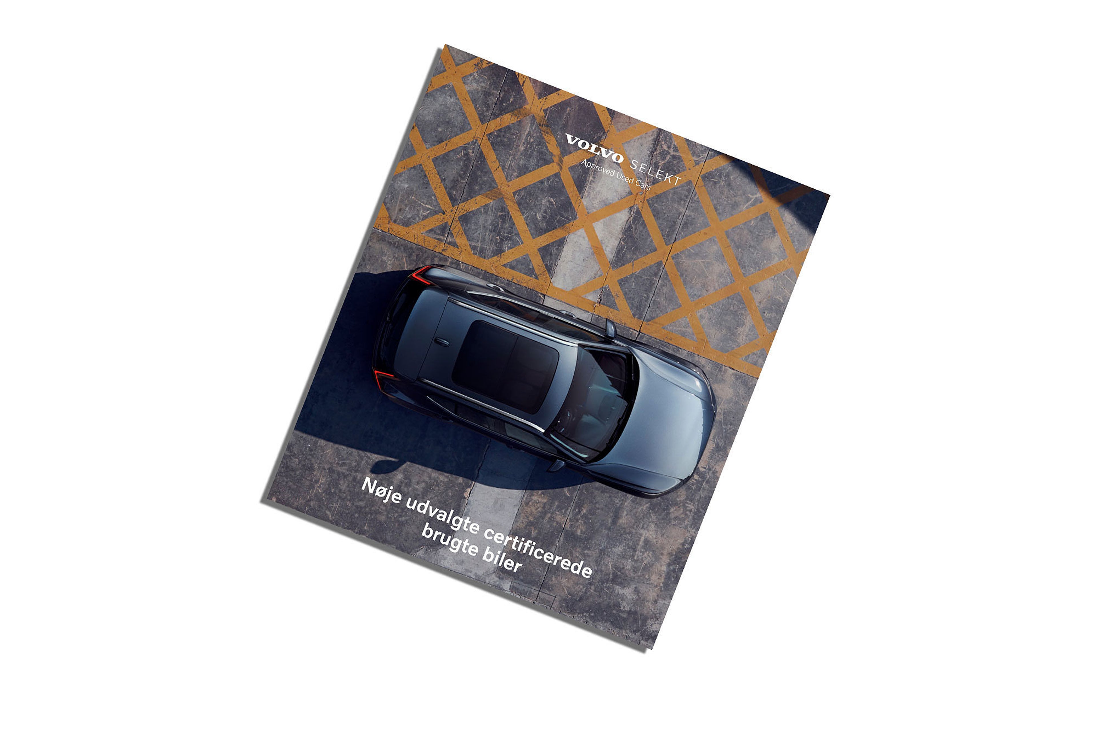 Topvinkel af en mørkegrå Volvo-bil parkeret på en betonoverflade med gule diagonale linjer. Teksten "Volvo Selekt: Godkendte Brugte Biler" er synlig.