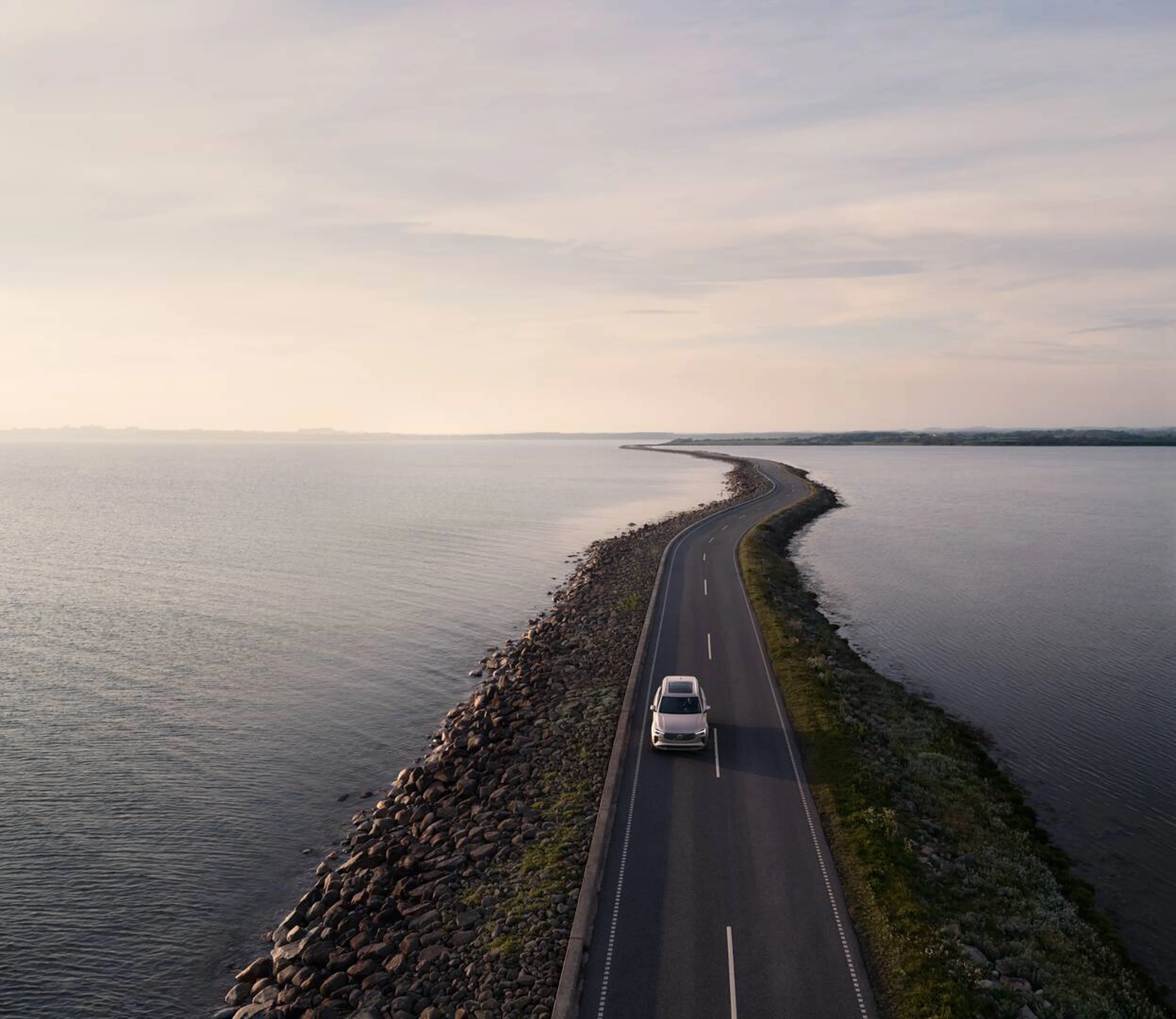 Luftfoto af en Volvo bil, der kører på en smal vej omgivet af roligt vand på begge sider. Himlen er overskyet, hvilket skaber en fredfyldt og rolig stemning.
