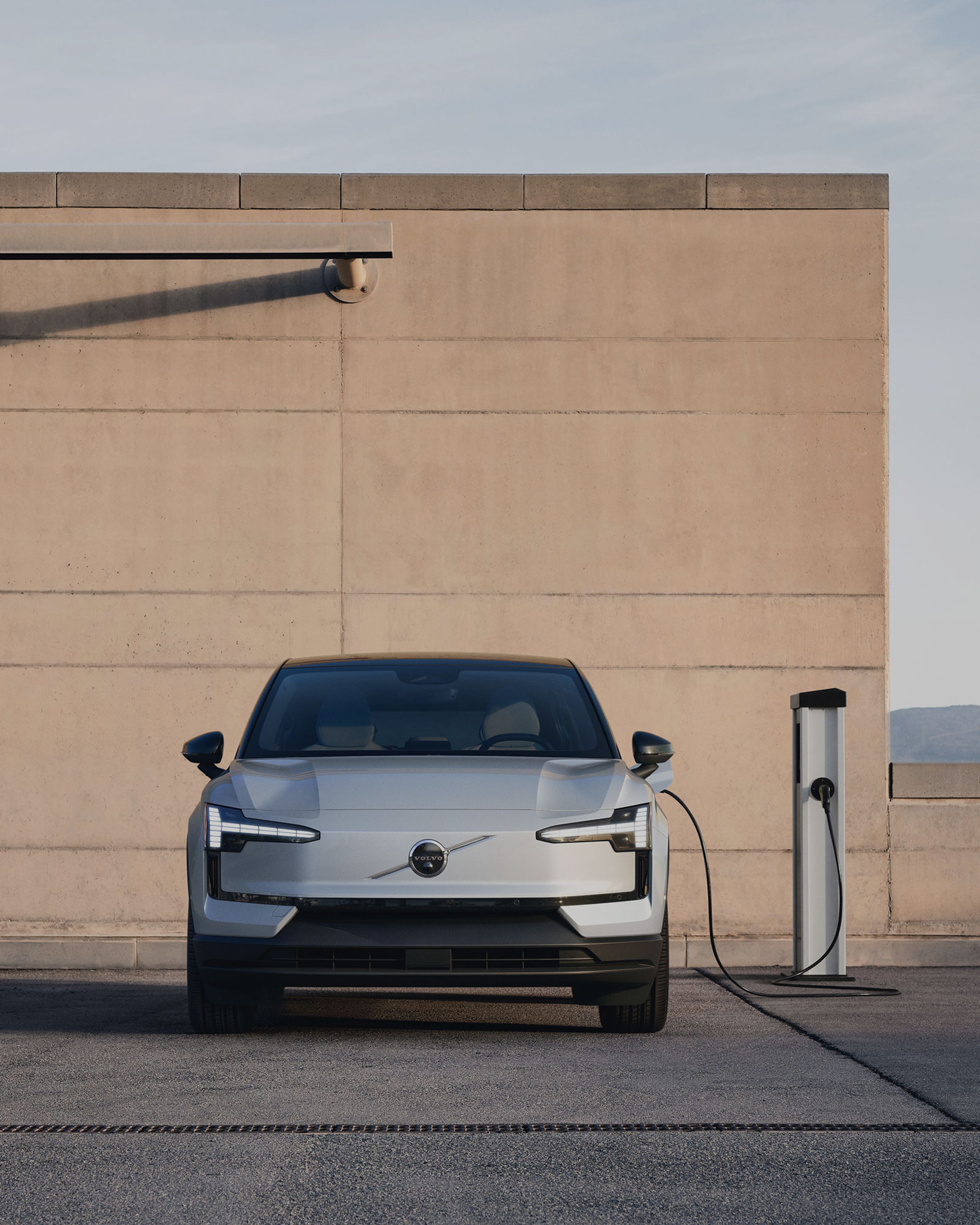 Voorkant van een grijze Volvo EX30 met zwart dak gekoppeld aan een laadpaal op een parking buiten.