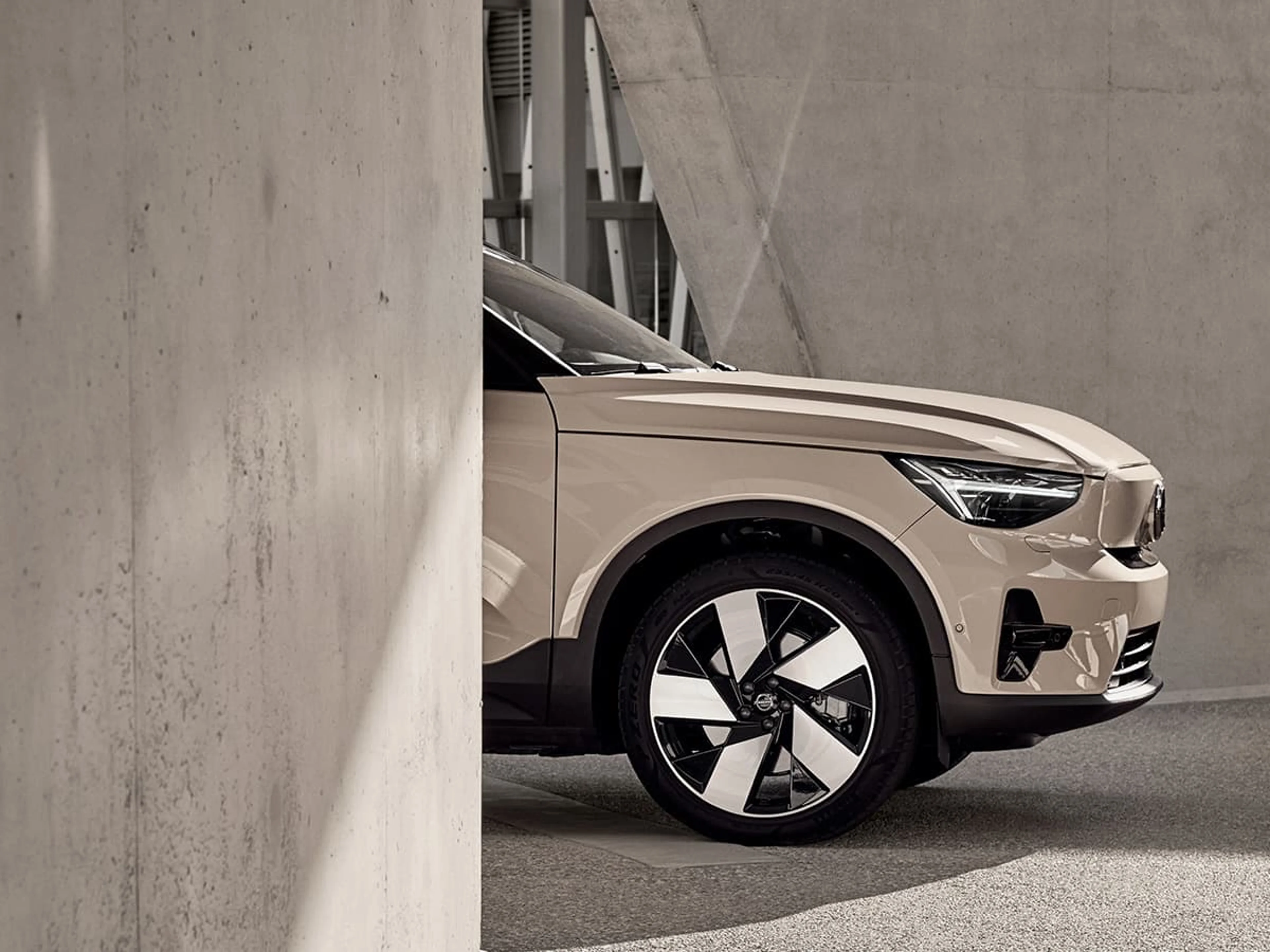 Gros plan sur l'avant d'une Volvo EX40 de couleur beige, partiellement visible derrière un mur en béton dans un décor urbain.