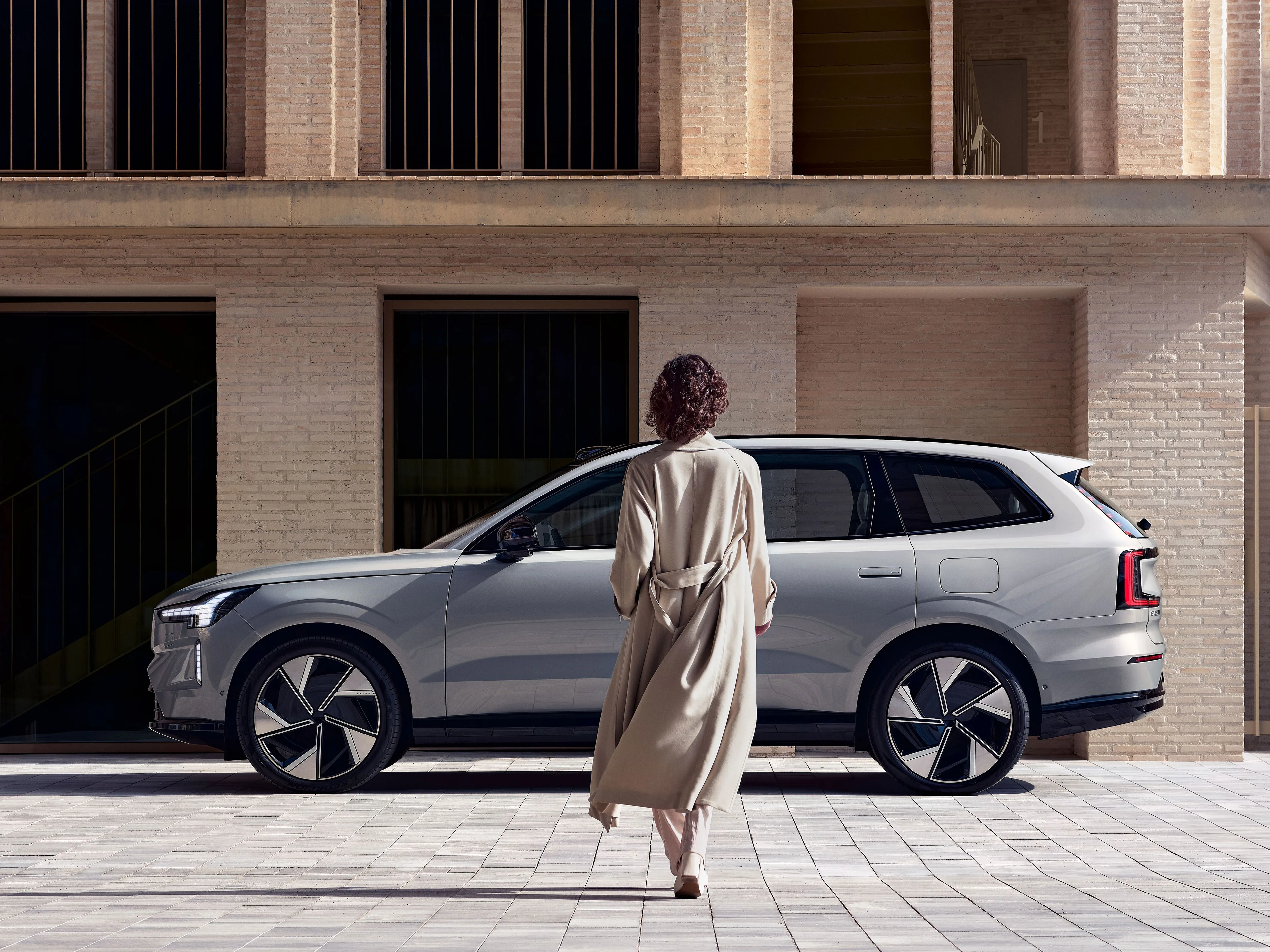 Eine Frau im hellen Trenchcoat schreitet auf einen silbernen Volvo EX90 zu.