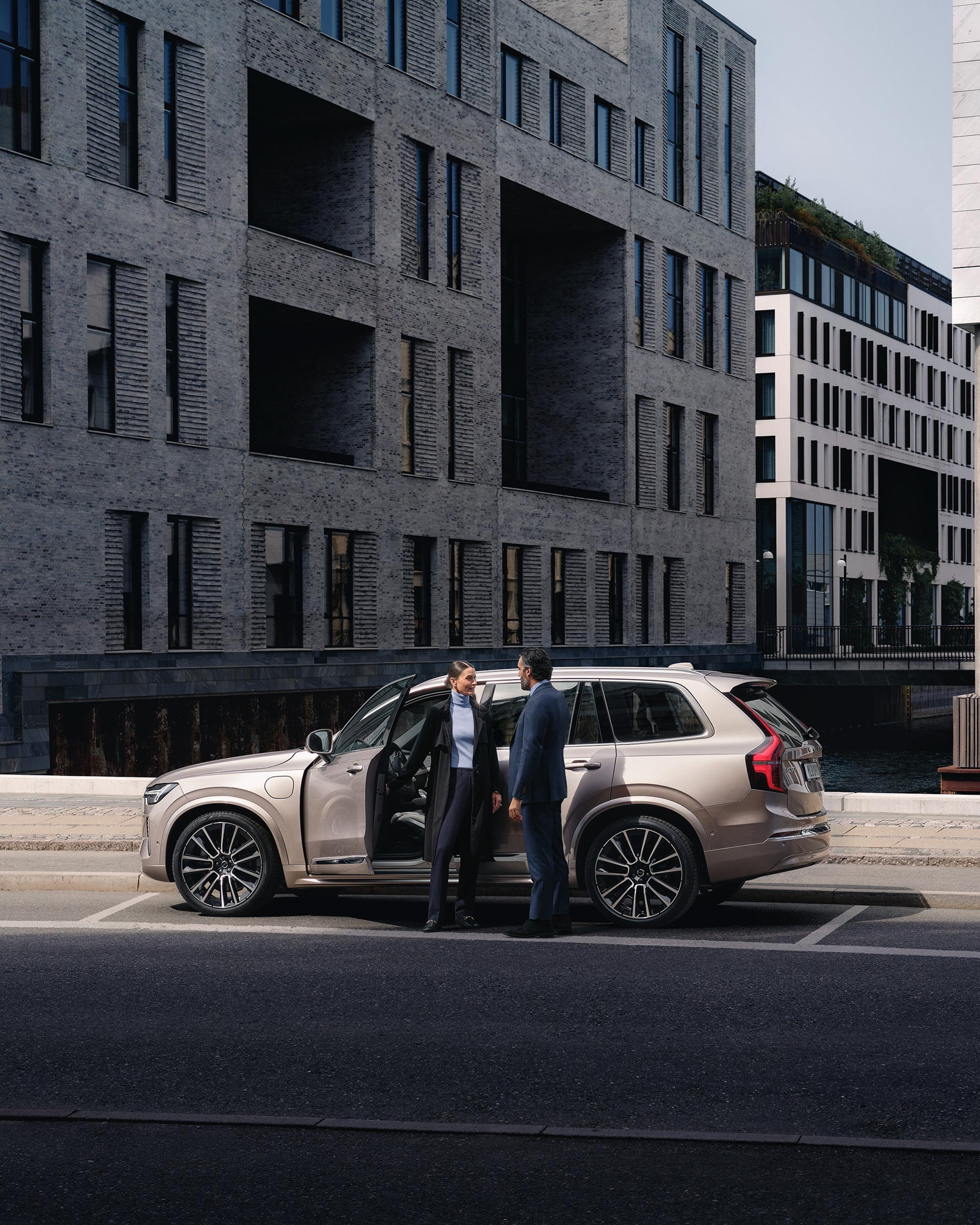 Nainen ja mies keskustelevat kadun varteen pysäköidyn Bright Dusk -värisen Volvo XC90 lataushybridikatumaasturin edessä.