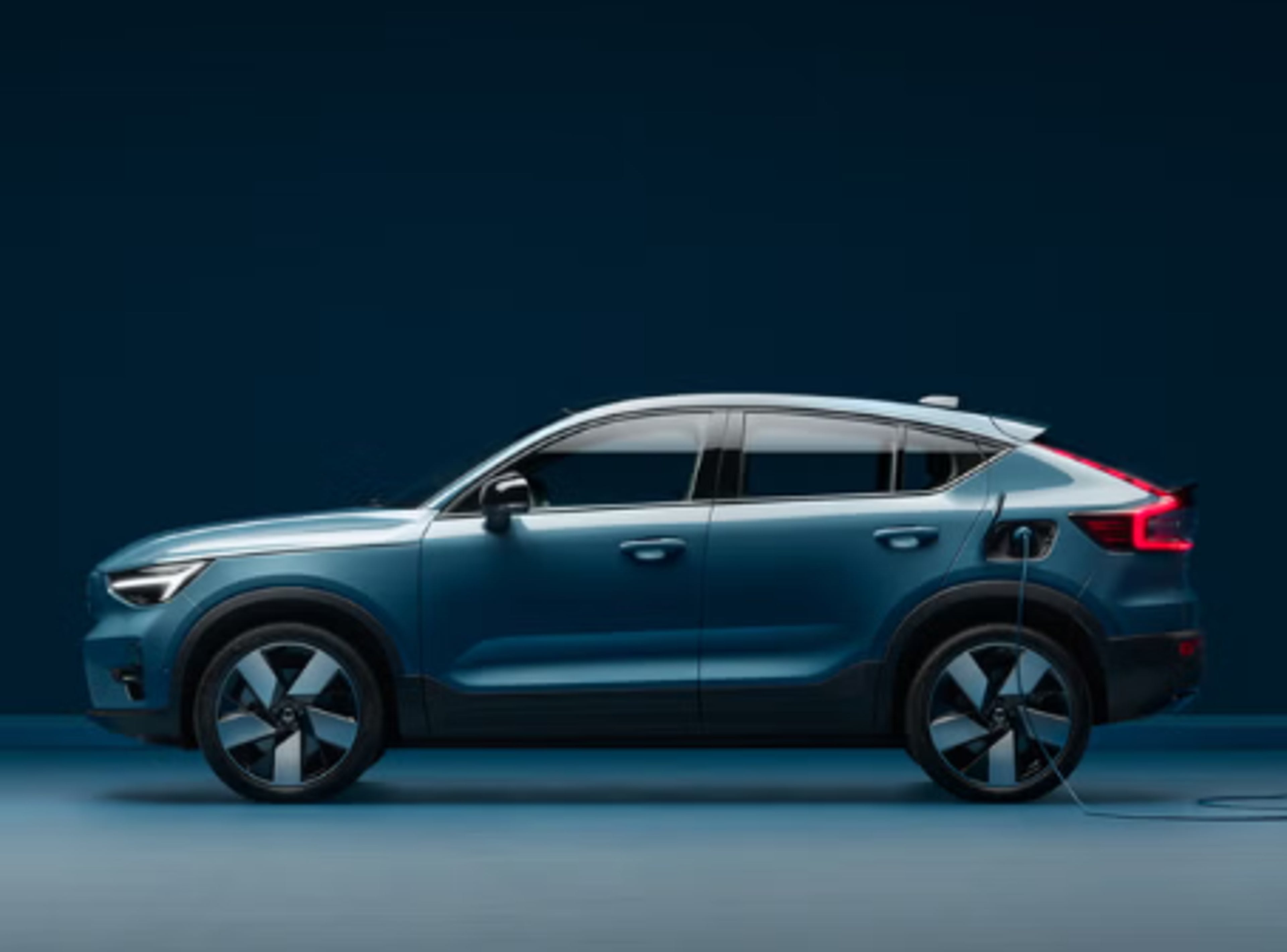 En elegant blå elektrisk Volvo C40 bil er tilsluttet en oplader foran en mørkeblå baggrund. Bilens moderne design og glatte linjer udstråler innovation og bæredygtighed.