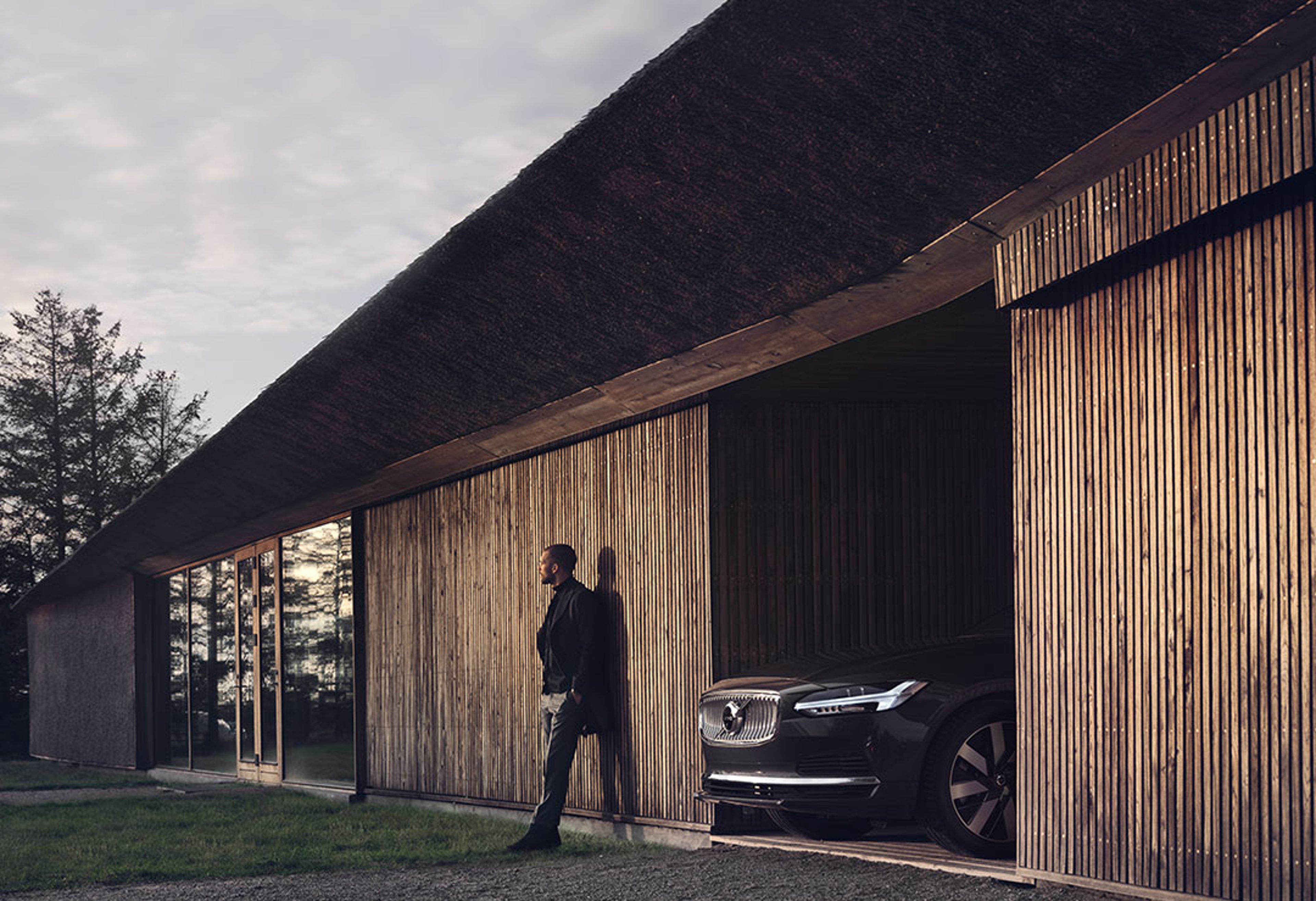 Ein dunkelgekleideter Mann lehnt mit dem Rücken zur Wand neben einer geöffneten Garage an einem holzvertäfelten Wohngebäude. Aus der Garage ragt die Front eines schwarzen Volvo V90.